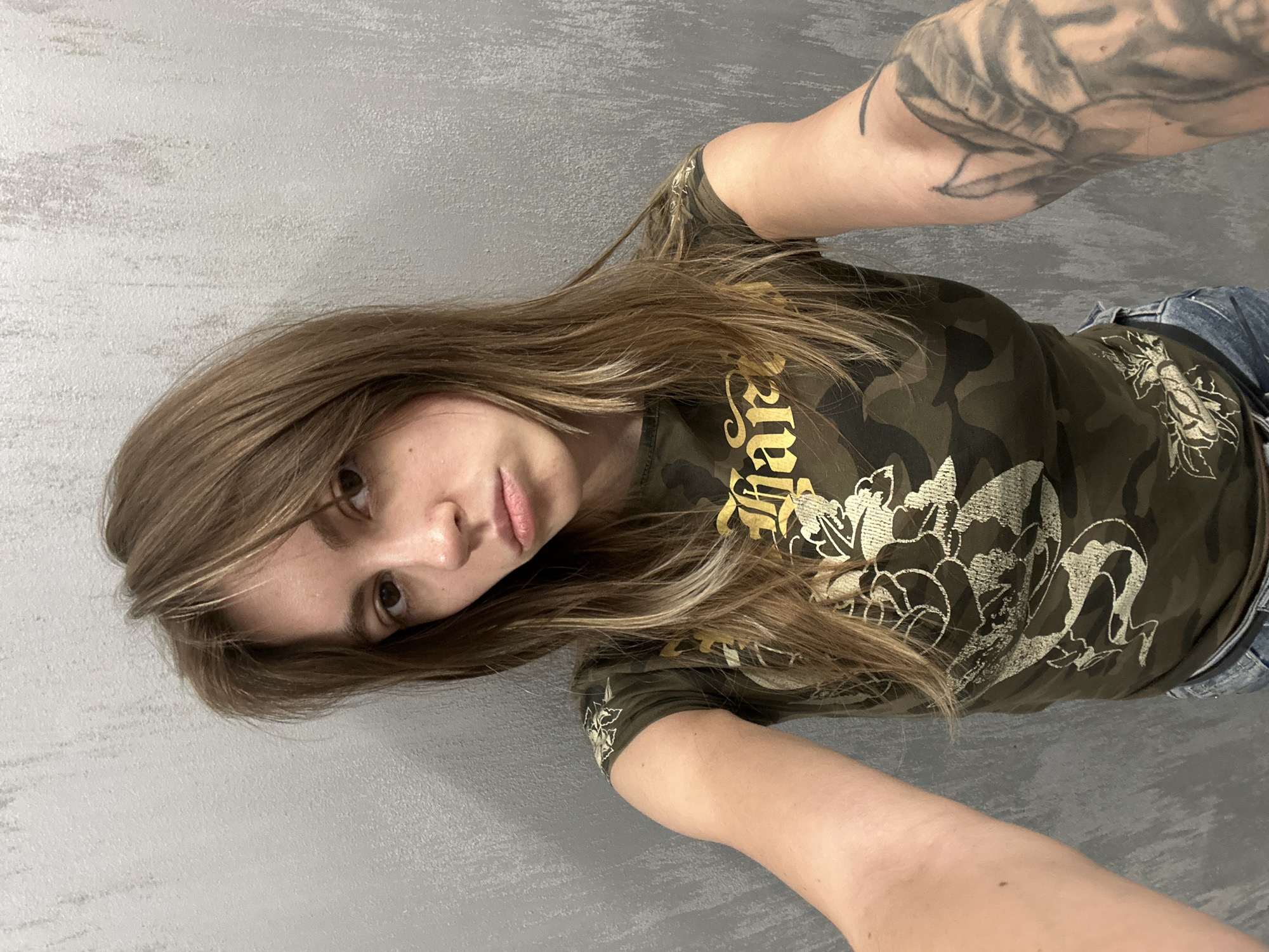 A young woman with shoulder-length brown hair and a neutral expression is taking a selfie. She is wearing a camouflage t-shirt with a gold graphic design on it. The background is a gray, textured wall. The image appears to have been taken indoors, possibly in an urban setting like Moscow, Russia.