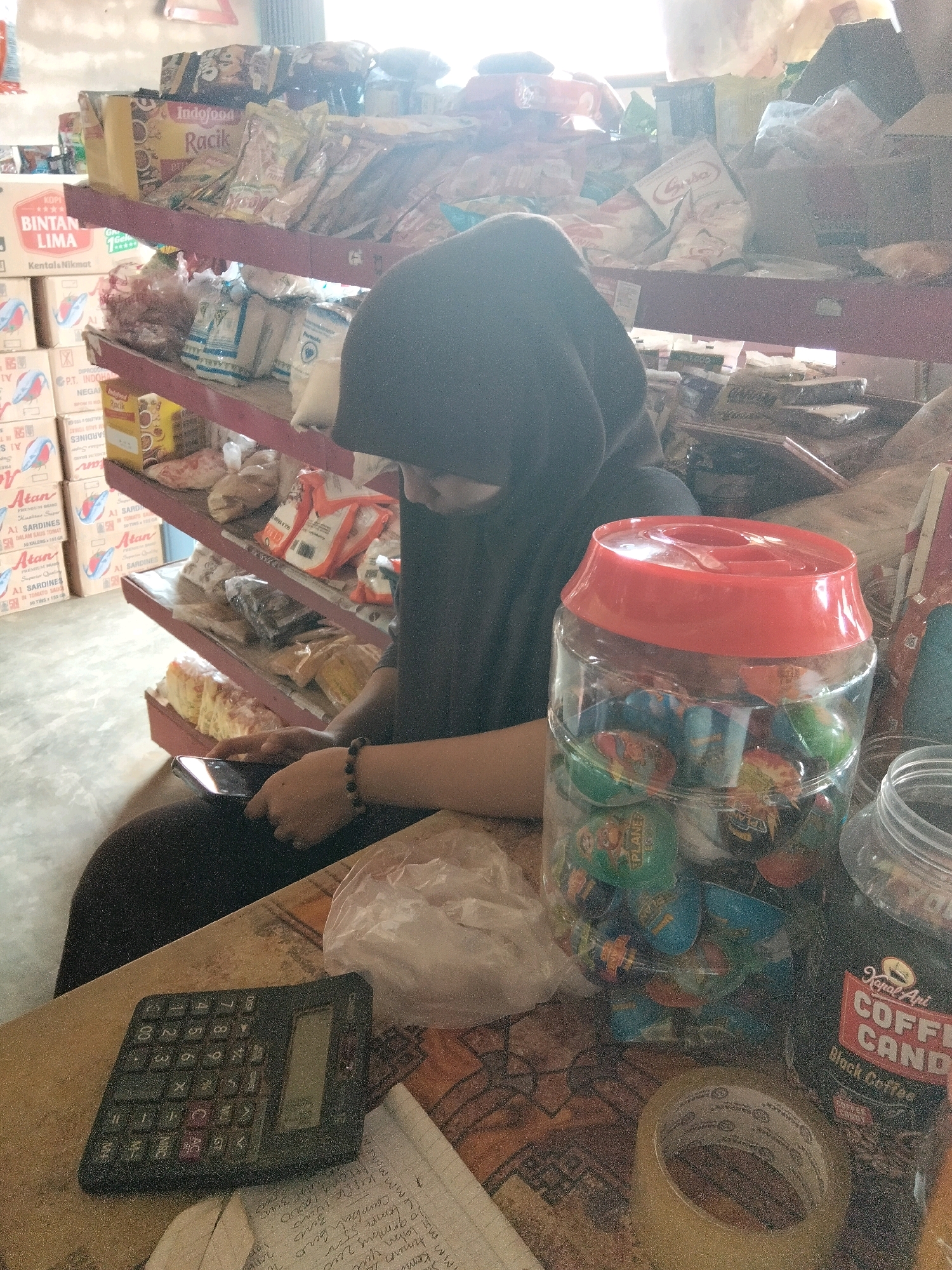 The image depicts a person wearing a dark hijab and a black top, seated in what appears to be a small convenience store or warung in Sidomulyo, Indonesia. The person is looking down at a smartphone held in their left hand, with their right hand resting on the phone. A black calculator is positioned on a wooden surface in front of them, along with a roll of clear tape and a partially filled plastic container holding colorful, round items, possibly candies or toys. Behind the person, shelves are filled with various packaged goods, including food items and drinks. The store environment is dimly lit, with sunlight filtering in from the right, creating a bright haze. The overall impression is one of a casual moment, possibly during business hours, where the individual is taking a break or engaging with their device amidst the products for sale.