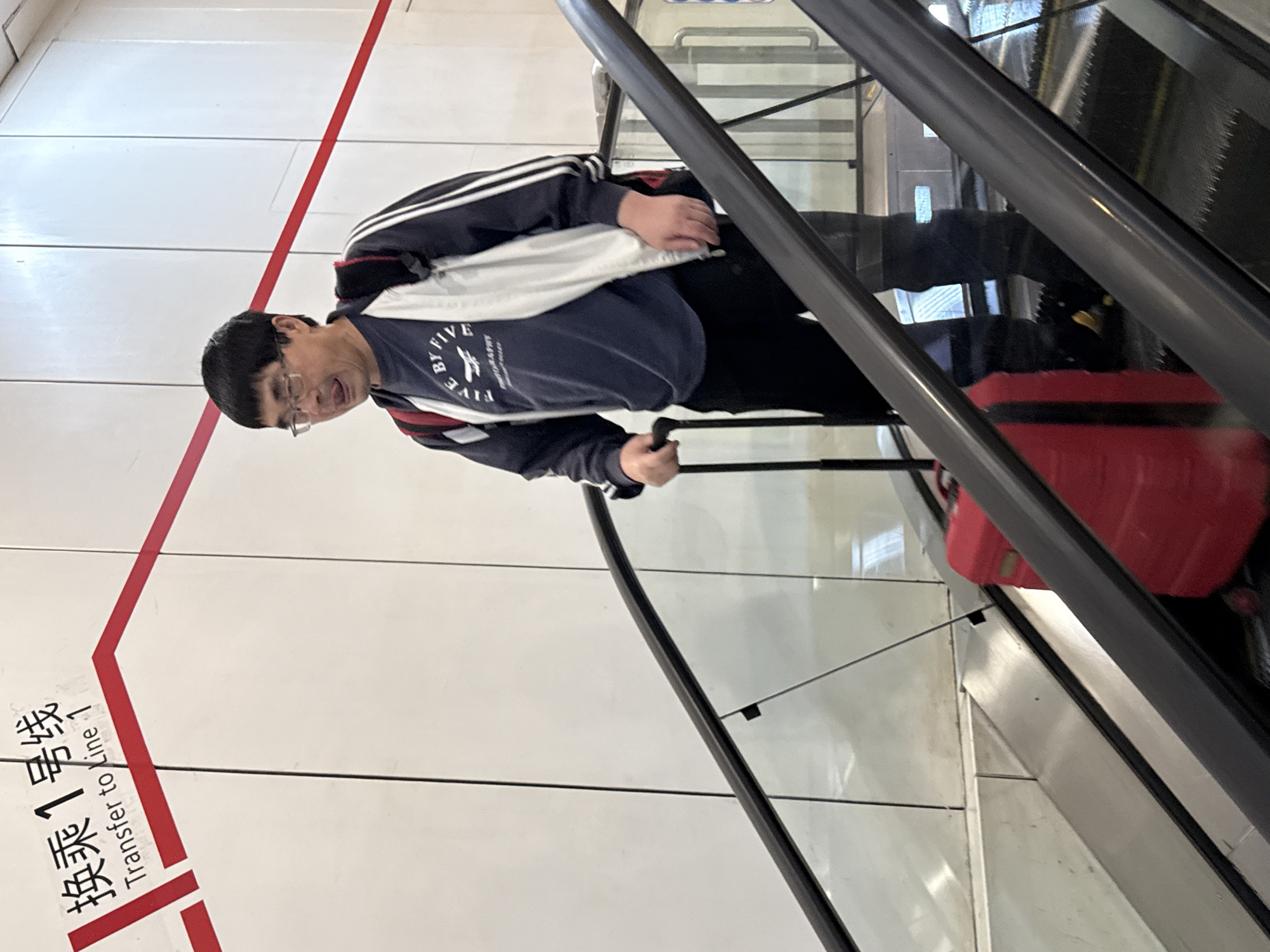 A man with short black hair and glasses is riding an escalator with a red suitcase in what appears to be a subway station. The man is wearing a dark blue sweatshirt with white and red accents, black pants, and a white bag slung over his shoulder. He is holding onto the escalator handrail. The escalator has a glass safety barrier.
To the left of the escalator is a white wall with a red stripe and the text "换乘 1 号线 Transfer to Line 1" in Chinese and English. The setting suggests the location is within a transportation hub in a Chinese-speaking region. Given the request specified Yuzhong District, China, it can be inferred that the photo was taken within a subway station of that location.