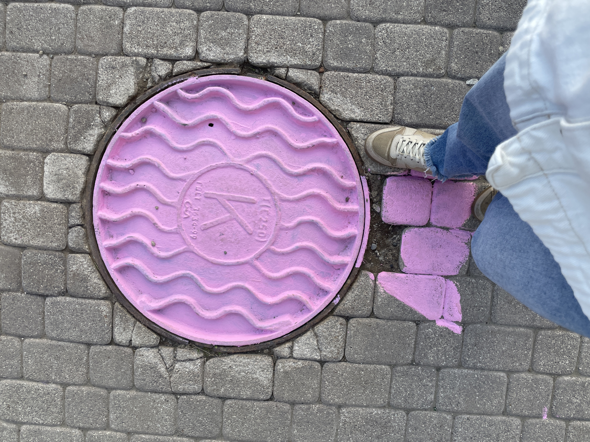 A top-down shot shows a pink-painted manhole cover, a person's legs and feet, and a paved area, likely a sidewalk. The manhole cover is round, with a raised wave pattern and a prominent "K" design in the center. Text is visible around the design. The manhole sits within a paving of gray rectangular blocks. A person, wearing sneakers, blue jeans, and a white top, is standing beside the manhole. Some of the blocks around the manhole are also painted pink, seemingly matching the manhole's color. The location is Izhevsk, Russia.
