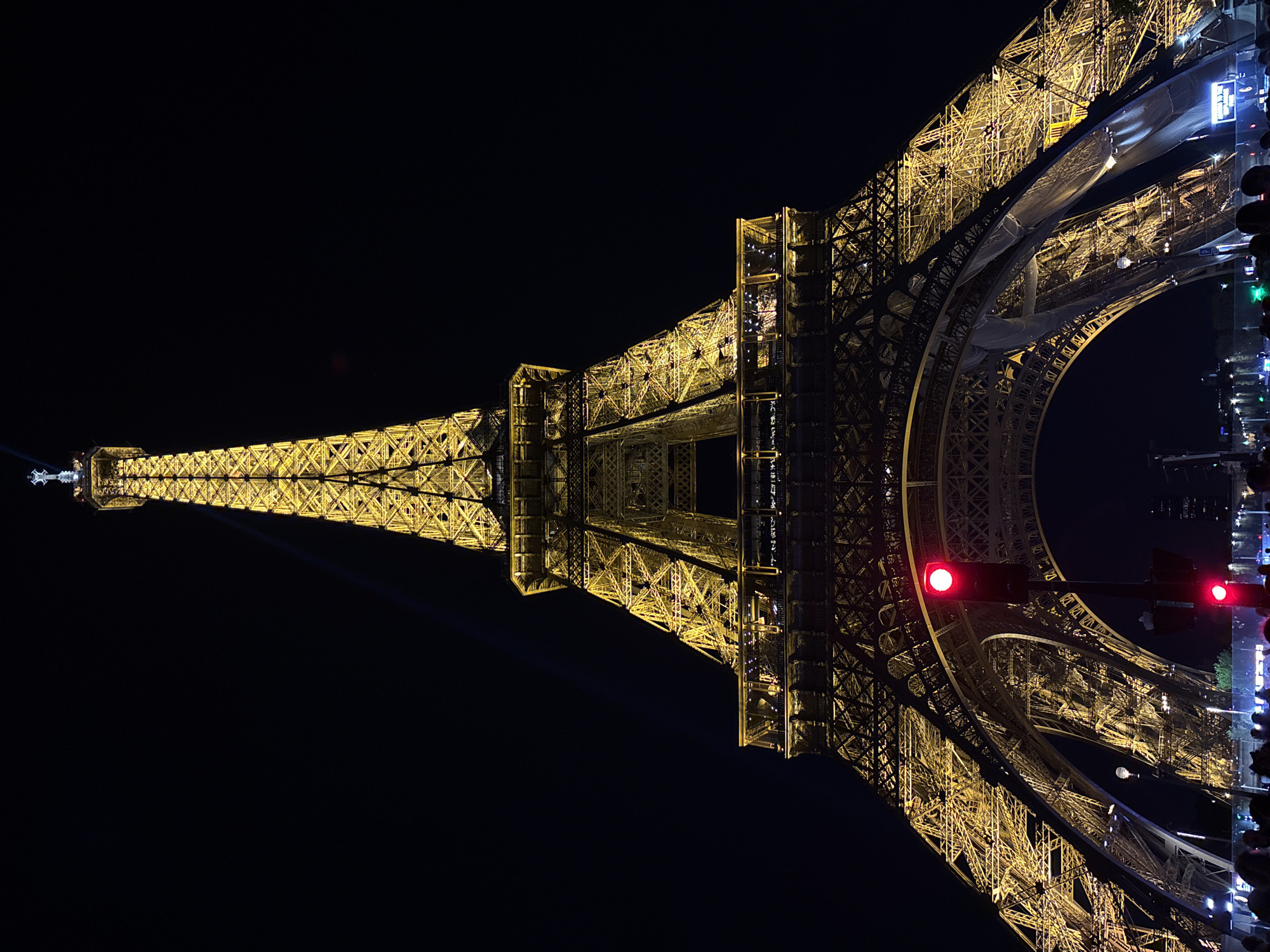 The image depicts the Eiffel Tower at night, illuminated in yellow light against a dark sky. The tower's metal framework is clearly visible, and the top portion is slightly pointed towards the left. At the base of the tower, there are streetlights and traffic signals, including a red light. Some structures are partially visible near the base of the tower along with what looks to be the side of a building. The sky is dark. The image is rotated counter-clockwise.