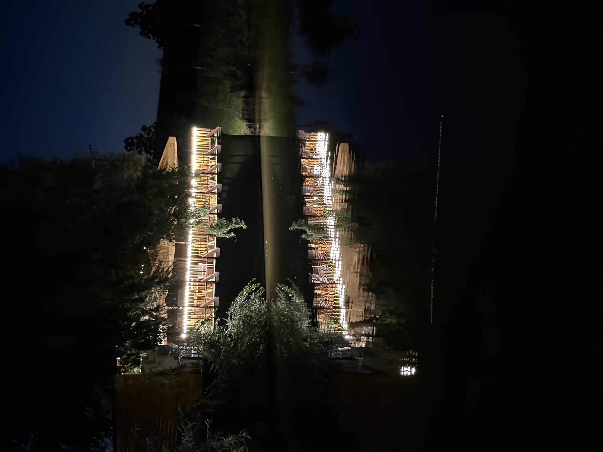 The image depicts a wooden structure, possibly a restaurant or a viewing platform, illuminated by string lights, situated on the edge of a body of water at night. The building has a balcony or deck with a railing, also lit by the lights. Lush green foliage partially obscures the structure. The water in the foreground reflects the lights and the building, creating a symmetrical, albeit distorted, image on its surface. The background is dark, with the silhouette of trees and a faint outline of a hill against a deep blue, night sky. There are no people visible in the image. The scene appears to be in a natural or semi-natural setting, likely on the outskirts of a town or city. This location is identified as being in the city of Strășeni, Moldova.