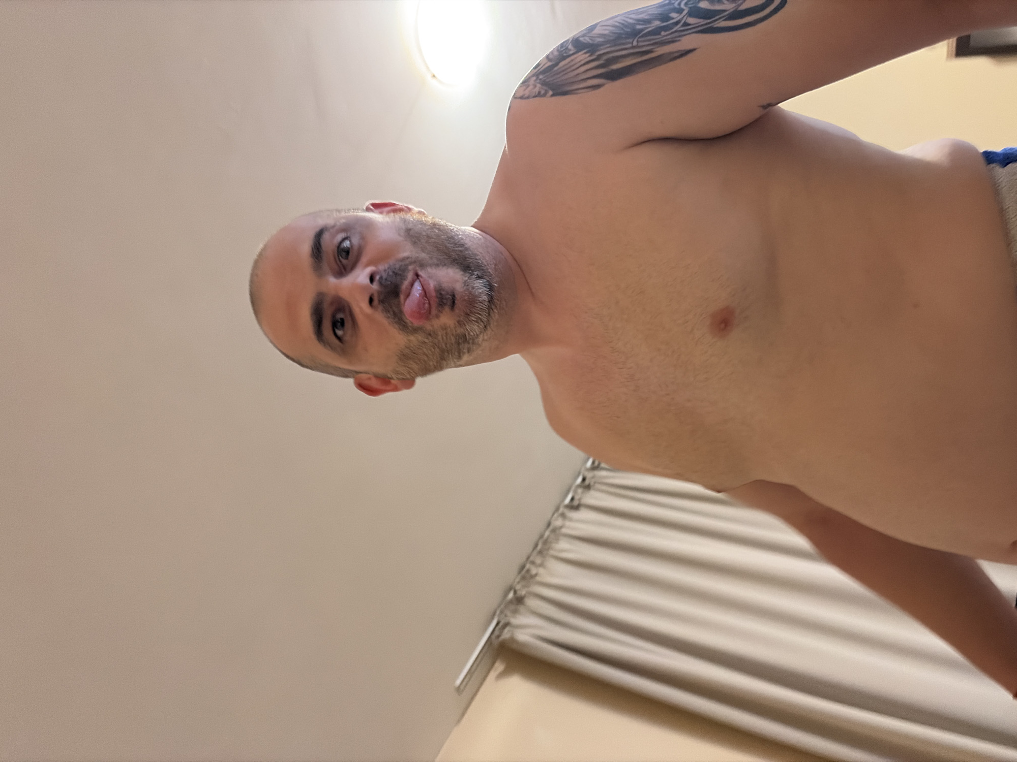 A low-angle, medium shot captures a shirtless man with a bald head, goatee, and wide eyes sticking his tongue out. He has a tattoo of wings on his left shoulder. The man is indoors, with light-colored curtains in the background. The setting appears to be a room in a residential building in the city of Nesebar, Bulgaria.
