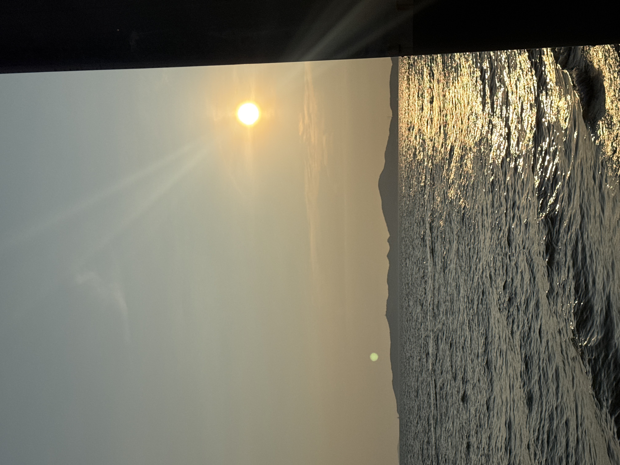 The image is a view of the sea in Pattaya City, Thailand, taken at sunset. The sun is high in the sky, emitting bright rays. The sea surface is textured with small waves reflecting the sunlight, creating a shimmering effect. A dark landmass is visible in the background, likely part of the coastline. The image appears to be taken from a high vantage point, with a portion of a dark structure visible at the top edge of the frame, possibly a building or platform. The overall scene depicts a tranquil seaside setting at dusk.