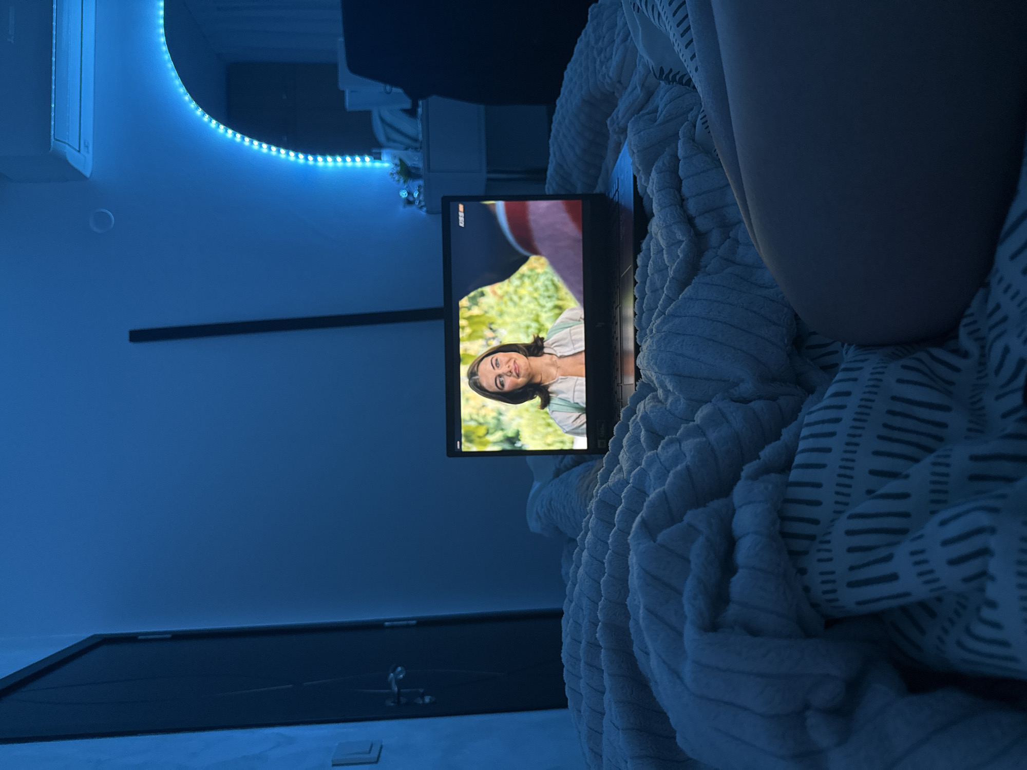 The image shows a person lying in bed, covered in a textured, patterned blanket, with a laptop resting on the blanket. The laptop screen displays a woman with shoulder-length brown hair smiling at the camera, set against a background of greenery. The room is illuminated with blue LED lighting, highlighting the air conditioning unit and a mirror on the wall. A dark door is visible in the background. The location is not discernible from the provided image, and it is impossible to determine if it is in Blagoevgrad, Bulgaria.