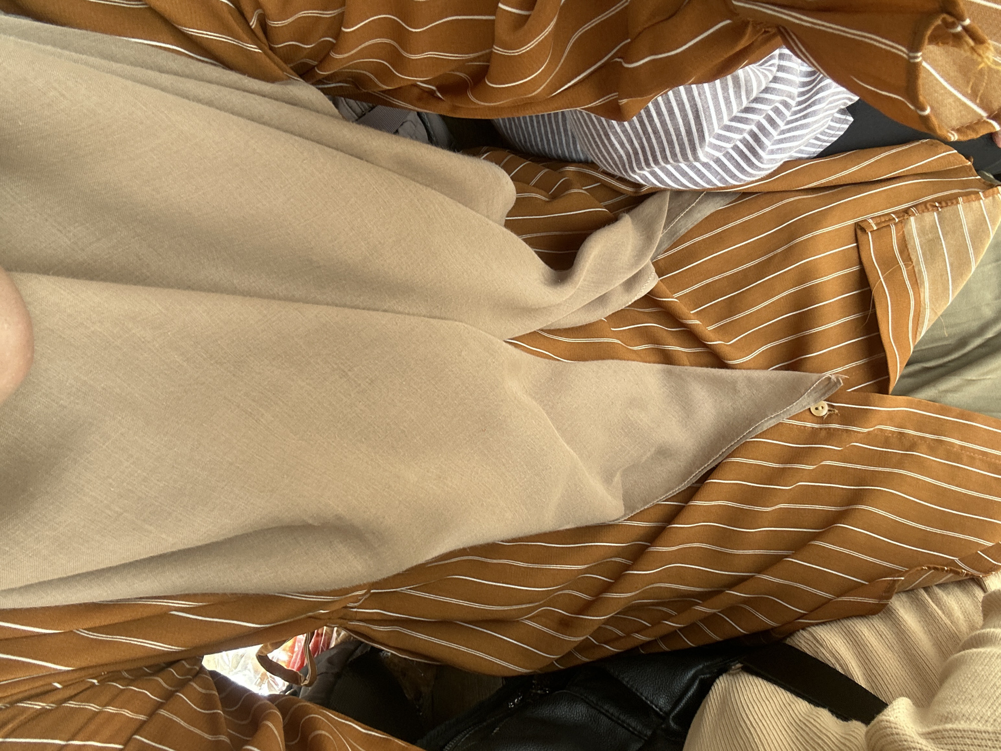 The image presents a close-up, top-down view of layered fabrics and clothing items, possibly an outfit being worn or arranged. The dominant colors are earthy tones, including light beige/tan, brown, and grey.
In the foreground and covering a large portion of the left side is a smooth, light beige or tan fabric, which appears to be draped and soft, possibly a headscarf, shawl, or a loose-fitting garment. Beneath and interspersed with this fabric is a rich brown fabric featuring thin, horizontal white stripes. This striped fabric shows a small, light-colored button in one section, suggesting it is part of a shirt or a dress.
Further in the background and partially obscured, a grey fabric with finer white stripes is visible. Towards the bottom right, a beige ribbed fabric, possibly a sweater or an undershirt, adds another texture to the composition. A black object, possibly a bag or part of another garment, is also seen amidst the clothing. A small portion of what appears to be skin, likely an arm or leg, is visible in the upper left corner, implying the clothes are being worn or handled.
The overall impression is one of textiles with varying textures and patterns, layered together. The lighting is soft and even, suggesting an indoor setting. There is no visible text in the image. The location context provided is Reuleut Timu, Indonesia.