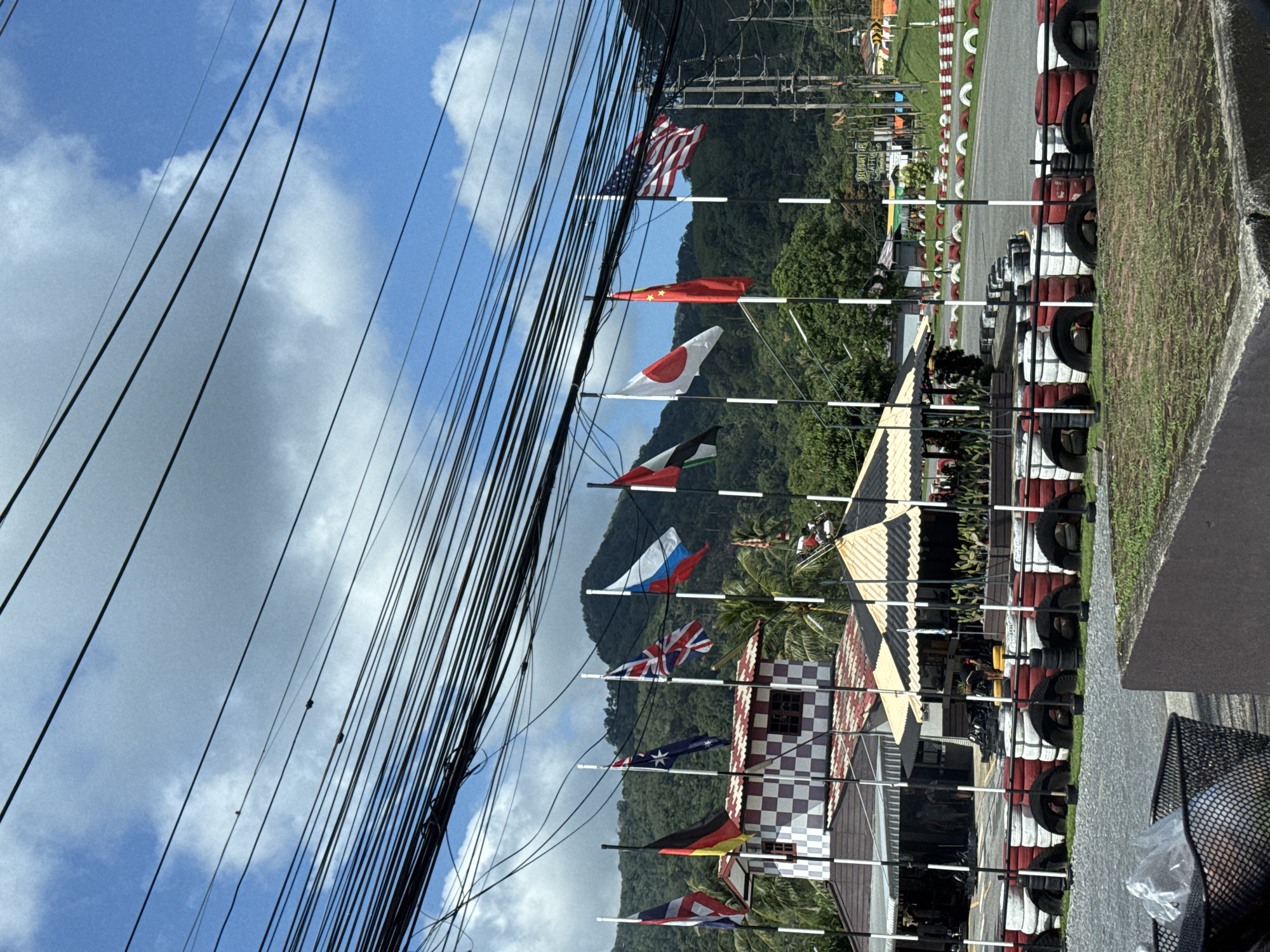 The image depicts an outdoor go-kart track in Kathu, Thailand, under a partly cloudy sky. A row of international flags flies from poles in front of a checkered building and a striped canopy structure. The flags visible include those of Thailand, Germany, Australia, the United Kingdom, Russia, the United Arab Emirates, Japan, China, and the United States. The track itself is bordered by tires stacked and painted red and white, with a gravel area in the foreground. Power lines crisscross the upper portion of the image against the sky, and lush green mountains form the backdrop. The scene appears to be a recreational area, possibly a tourist attraction, with no specific individuals or activities prominently featured.