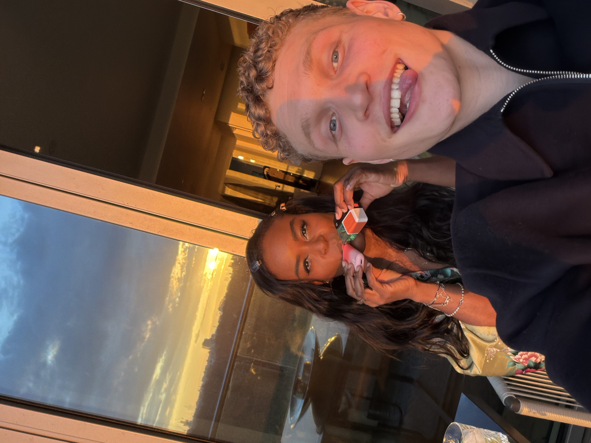 Two people are posing in front of a window with a view. The person in the upper portion of the frame is a fair-skinned individual with curly blonde hair, blue eyes, and a wide smile with their tongue slightly sticking out. They are wearing a dark zippered jacket.
Below them, a dark-skinned individual with long, dark hair is holding a small cube in their hands, with colors visible on its sides. They are wearing bracelets and are looking towards the camera.
Outside the window, the sky shows a sunset or sunrise with a mix of blue and orange hues, with what appears to be a cityscape visible in the orange glow. Given the time of the photo, the weather is nice. The location is suggested to be London, United Kingdom based on the backdrop.
