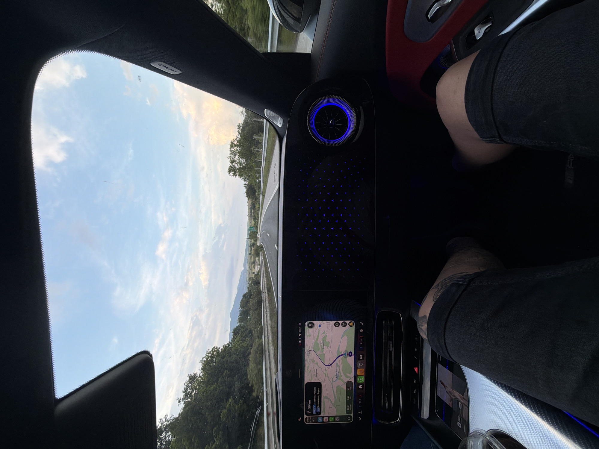 The scene is captured from the passenger seat of a high-performance Mercedes-Benz vehicle, likely an AMG model, as indicated by interior design elements and branding. The car is traveling at 92 km/h on a multi-lane highway in Bulgaria, specifically in the vicinity of Osikovitsa. The time is 19:59, and the ambient temperature is 21.5°C.
The expansive digital dashboard dominates the view, with the left portion displaying a navigation map. The route is set towards Pleven (Плевен), with intermediate points including Долни Дъбник, Горна Митрополия, and Trastenik. The map indicates a remaining distance of 30 km to Pleven and a total of 231 km to the final destination, with an estimated arrival time of 22:43. A segment of the current highway is labeled "A3" on the map. The right portion of the dashboard is illuminated with a recurring pattern of blue Mercedes-Benz star logos. A circular air vent, also illuminated in blue, is prominently featured on the far right of the dashboard. Blue ambient lighting outlines the lower dashboard and extends to the right door panel, which features red upholstery and a speaker grille. The right A-pillar is black and inscribed with "AIRBAG." A sunroof is partially open above the windshield.
In the foreground, the tattooed legs of an individual, wearing black shorts, are visible from a first-person perspective. The left leg rests near a textured silver surface, possibly part of the center console or cup holder area. The right leg is positioned against the red door panel.
Outside the vehicle, the highway extends straight into the distance, bordered by metal guardrails and dense green foliage on either side. Beyond the immediate trees, rolling hills and mountains are visible under a partly cloudy sky, with light blue transitioning to soft white and pink hues, suggesting late afternoon or early evening light. The windshield exhibits minor dust or streaks.
