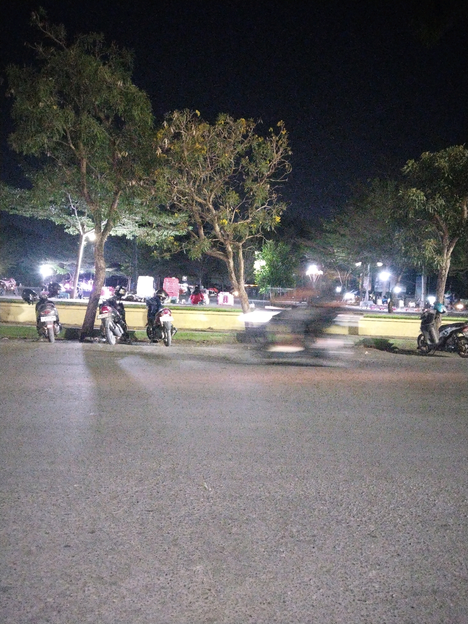 In Slawi, Indonesia, at night, a street scene unfolds with trees and parked motorcycles. The foreground is dominated by a paved road, with a slight upward incline towards the background. To the left, three motorcycles are parked along a yellow-painted curb, partially shaded by a tree. One motorcycle is in the immediate foreground, to the left of center, and two more are positioned to its right, closer to a tree trunk. The middle ground features a strip of grass behind the curb, and beyond that, several more motorcycles are parked. The background reveals a dimly lit area with scattered lights, suggesting buildings or stalls. Trees are prominent throughout the scene, with some illuminated from below, casting shadows. A blurred figure on a motorcycle is moving from left to right across the middle ground, indicating motion in the low-light conditions. The overall atmosphere is that of an outdoor public space at night.