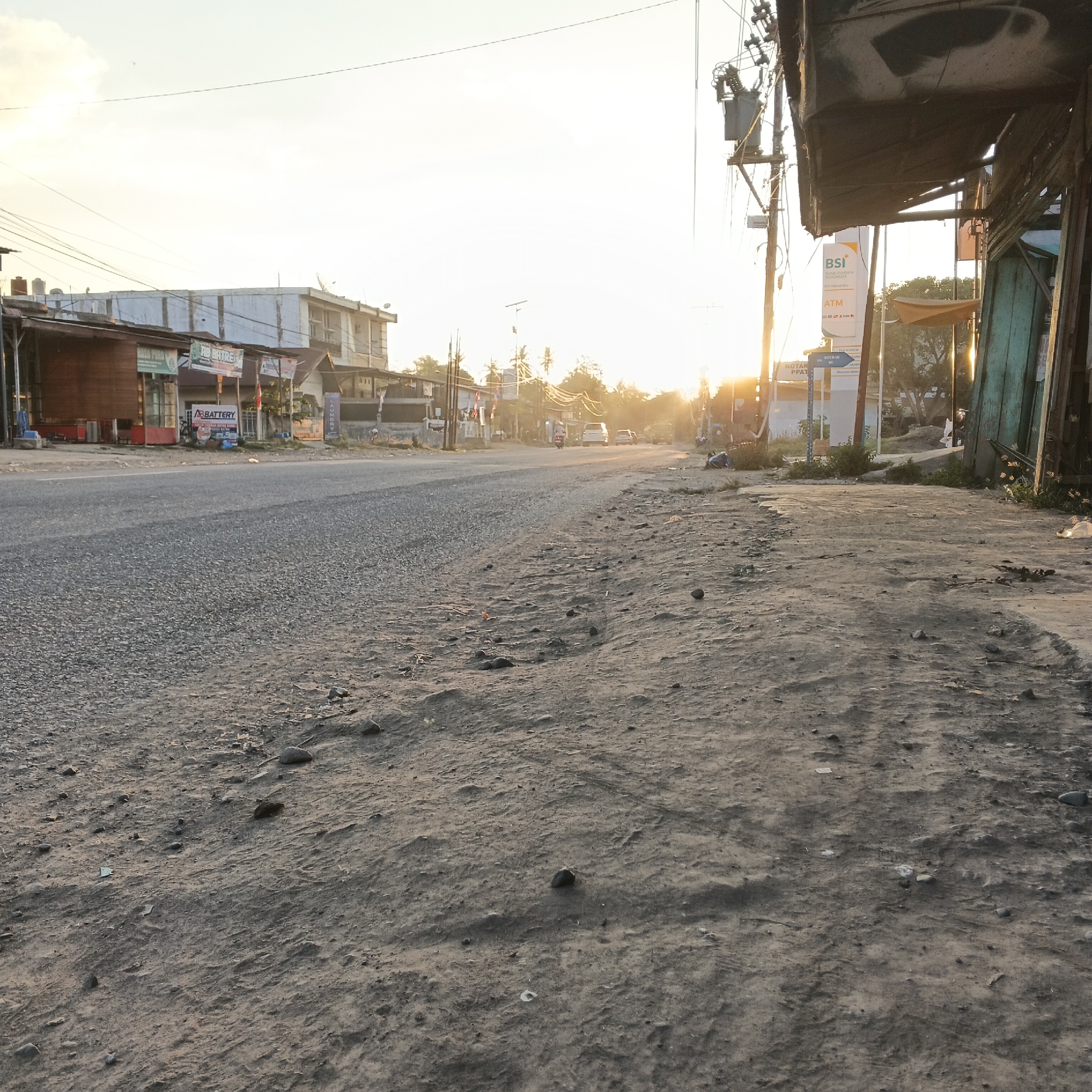 The image depicts a roadside scene in Meunasah Raya, Indonesia, during sunset. The foreground is dominated by a dusty, unpaved shoulder of the road, scattered with small stones and debris. A paved asphalt road stretches into the distance, lined with buildings and utility poles. The buildings appear to be a mix of shops and residences, some with visible signage. A few vehicles, including a motorcycle and cars, are present on the road. A person is visible sitting on the ground on the right side of the frame. The sky is bright with the setting sun casting a warm glow over the scene, highlighting the silhouette of trees and power lines. The overall atmosphere is that of a typical street in a developing area transitioning from day to evening.