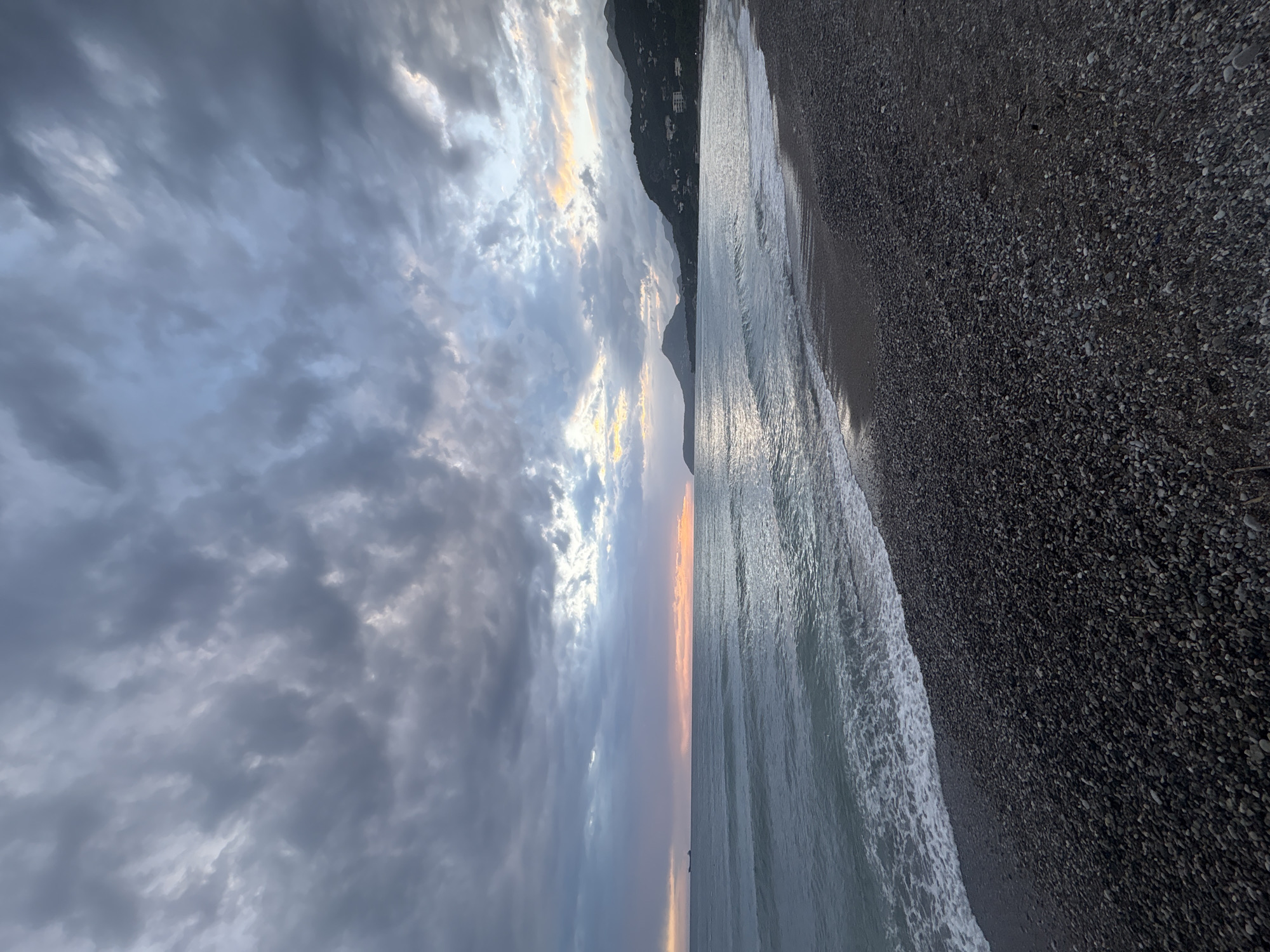 The image depicts a beach scene in Bar, Montenegro, at dusk or dawn. The composition is vertically oriented, with the shoreline bisecting the frame. To the right is a dark, pebbly beach sloping upwards out of view. The sea occupies the central portion of the image, with gentle waves lapping at the shore. The sky, on the left, is dominated by grey and blue clouds. There is a small silhouette of a mountain to the left. No people or animals are visible. The lighting suggests a sunrise or sunset, with faint orange hues peeking through the clouds near the horizon.