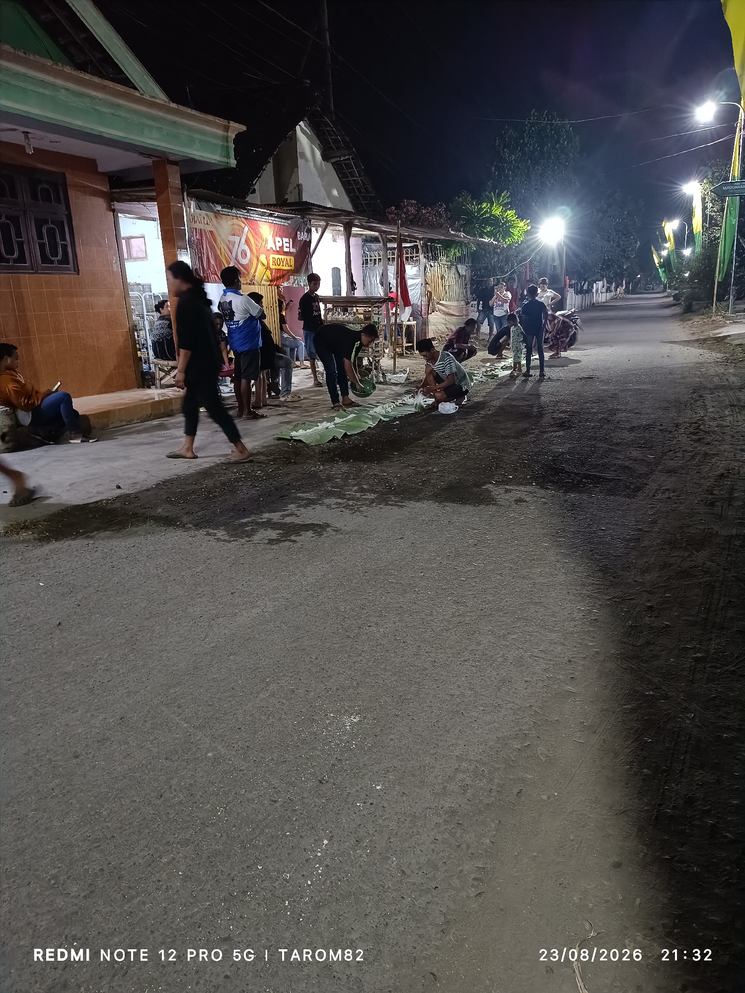 In Rejoagung, Indonesia, at night, a group of people are gathered on a paved street, illuminated by streetlights. In the foreground, several individuals are crouched down, some handling what appear to be food items laid out on large green leaves. The street is lined with buildings and trees, with banners and flags visible in the background, suggesting a festive occasion or a community event. Some individuals are standing, observing the scene or interacting with each other. The overall atmosphere is casual, with a sense of community gathering.