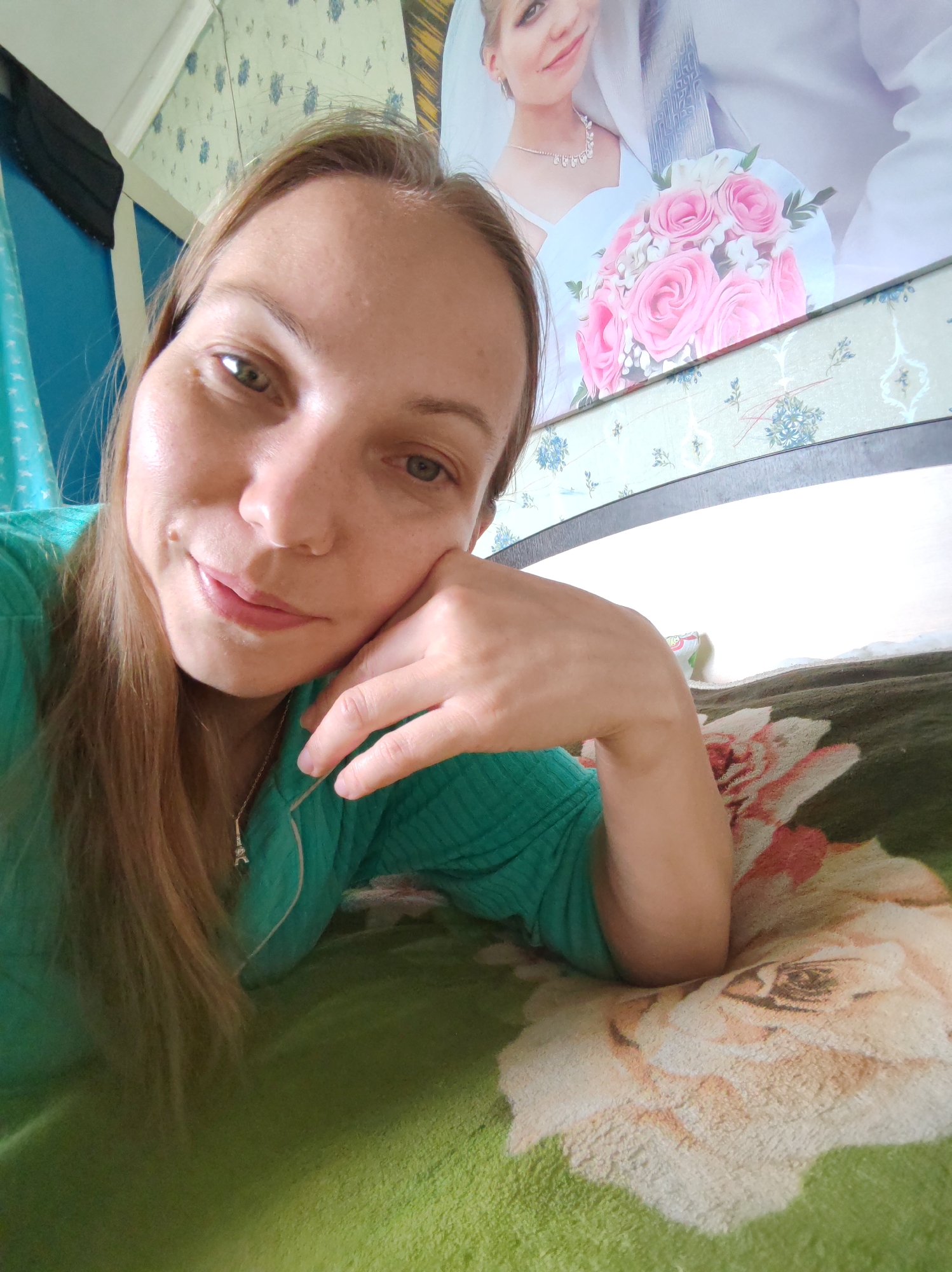 A woman with blonde hair and green eyes lies on a bed, her head propped on her hand. She is wearing a green shirt. In the background, a framed picture of a wedding is visible. The environment appears to be a bedroom in an apartment or house. The image is a selfie. No specific location in the city of Чухлэм, Russia can be determined from the image.