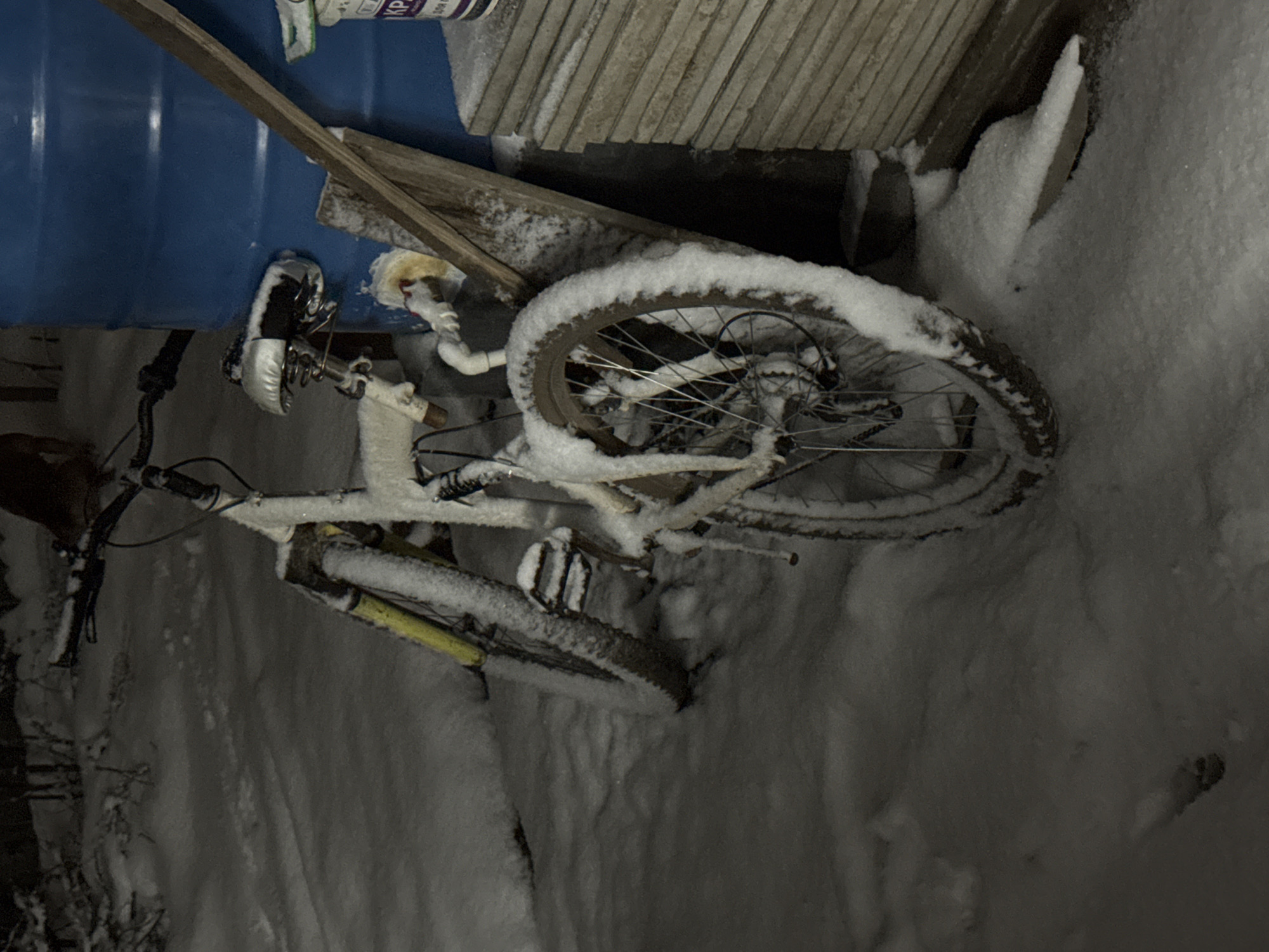In Chaikovsky, Russia, a bicycle covered in snow rests against a blue barrel and stacked gray slabs. The surrounding ground is also covered in snow.