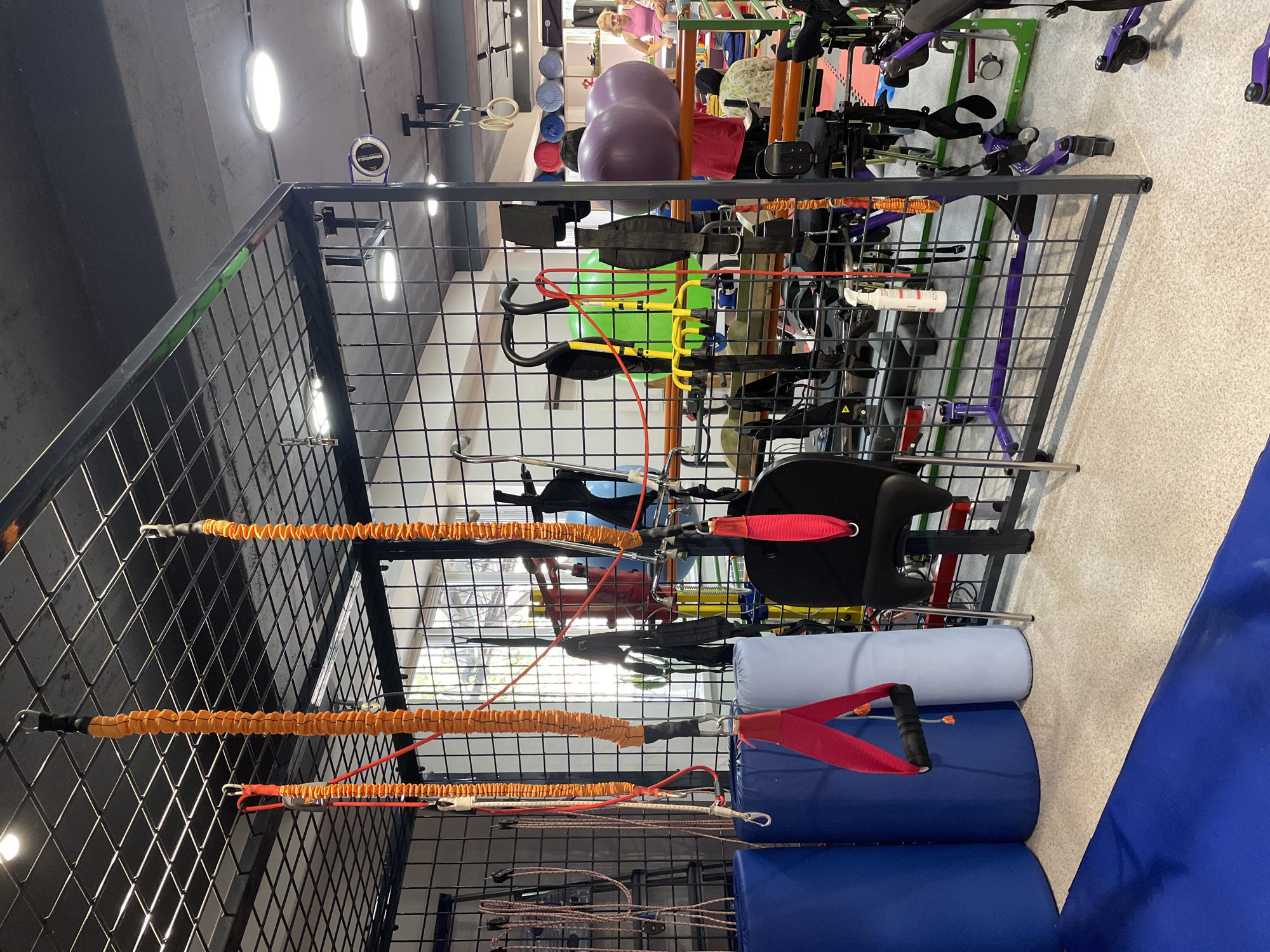 The image shows an indoor rehabilitation gym equipped for physical therapy. In the foreground, a black metal cage-like structure holds various exercise equipment. Suspended from the ceiling within the cage are orange resistance bands with black connectors and red handles, as well as other red straps. To the left, blue and light blue cylindrical bolsters are visible. A black chair with a red seat strap is also present inside the cage. Beyond the cage, the gym extends into a larger space. In the middle ground, there's a bright green exercise ball and yellow equipment with handles. Further back, large purple exercise balls are stacked. Several people are present in the background, including a woman in a pink top and a person wearing a headscarf. There are also specialized mobility aids, such as walkers with wheels, visible in the background. The environment appears well-lit with overhead fluorescent lighting. The flooring is a light-colored speckled material. This scene depicts a specialized facility likely dedicated to physical rehabilitation, possibly located in Almaty, Kazakhstan, given the context.