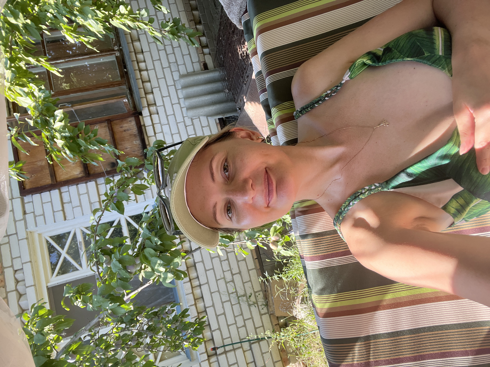 A woman with blonde hair, wearing a green baseball cap and a green and white patterned bikini top, is sitting on a striped outdoor swing. She is looking at the camera and smiling. Behind her is a white brick building with a window and a wooden shutter. There are green leaves and branches from a tree in the foreground, some of which have small purple fruits. The background shows a garden area with grass and weeds. The image appears to be taken on a sunny day.