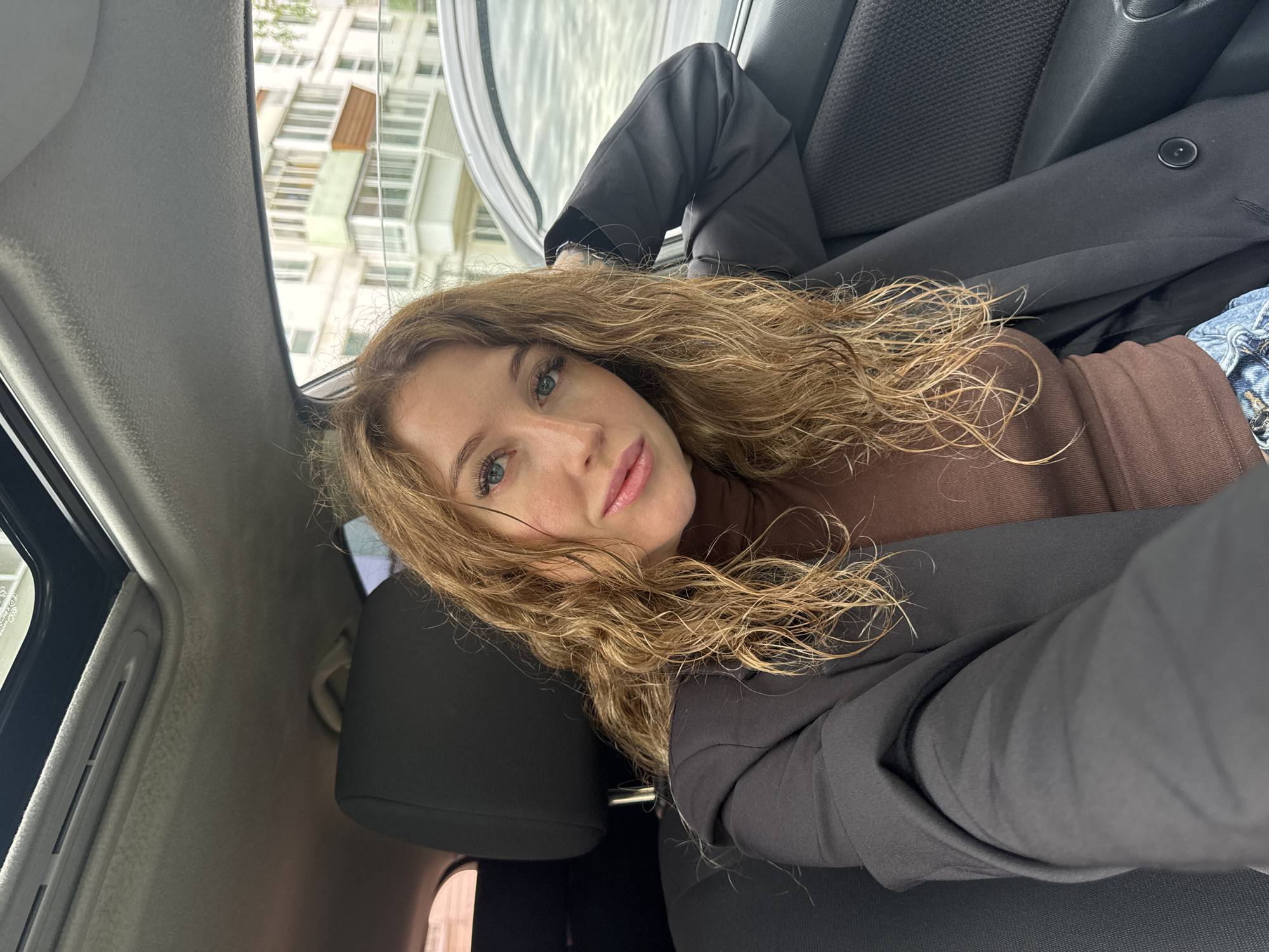 A woman with long, wavy blonde hair is sitting in the passenger seat of a car. She is wearing a brown turtleneck shirt and a black blazer. Her left arm is resting on the armrest. Outside the car window, a multi-story building with balconies is visible. The image appears to have been taken in Chaikovsky, Russia, based on the architectural style of the building and the overall environment.