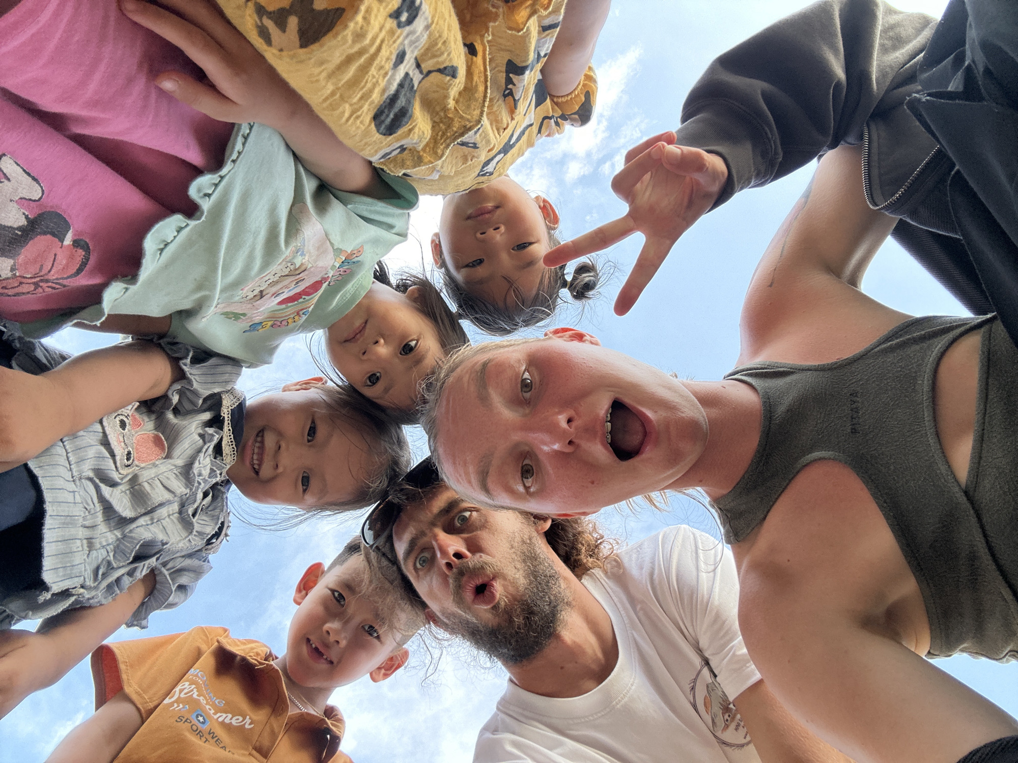 The image is a low-angle photograph taken outdoors in Gia Hội, Vietnam, depicting seven individuals looking directly down into the camera against a clear blue sky with minimal white clouds. The subjects are arranged in a circular formation, with their heads tilting inwards towards the lens.
In the immediate foreground, a woman with light-colored hair and visible dental braces has an open mouth and wide eyes. She wears a dark green sleeveless athletic top with a cut-out detail on the chest and holds her left hand up, forming a peace sign with her index and middle fingers extended. A dark, long-sleeved garment is partially visible on her right side.
To her left, a man with a beard and dark, curly hair presents a wide-eyed expression with his lips pursed into an 'o' shape. He wears a white t-shirt. Adjacent to the man, a child in an orange t-shirt with text visible on the shoulder looks down. Continuing clockwise around the circle, a child with dark hair wearing a blue and white striped top smiles faintly. Next, a child wearing a mint green t-shirt with a graphic design looks directly down. A fifth child, partially visible in a pink top with a Minnie Mouse graphic, also looks down. The seventh individual is a child with dark hair styled in pigtails, wearing a yellow patterned garment, and looking directly into the lens. The composition emphasizes the communal, downward gaze of the group against the expansive sky.
