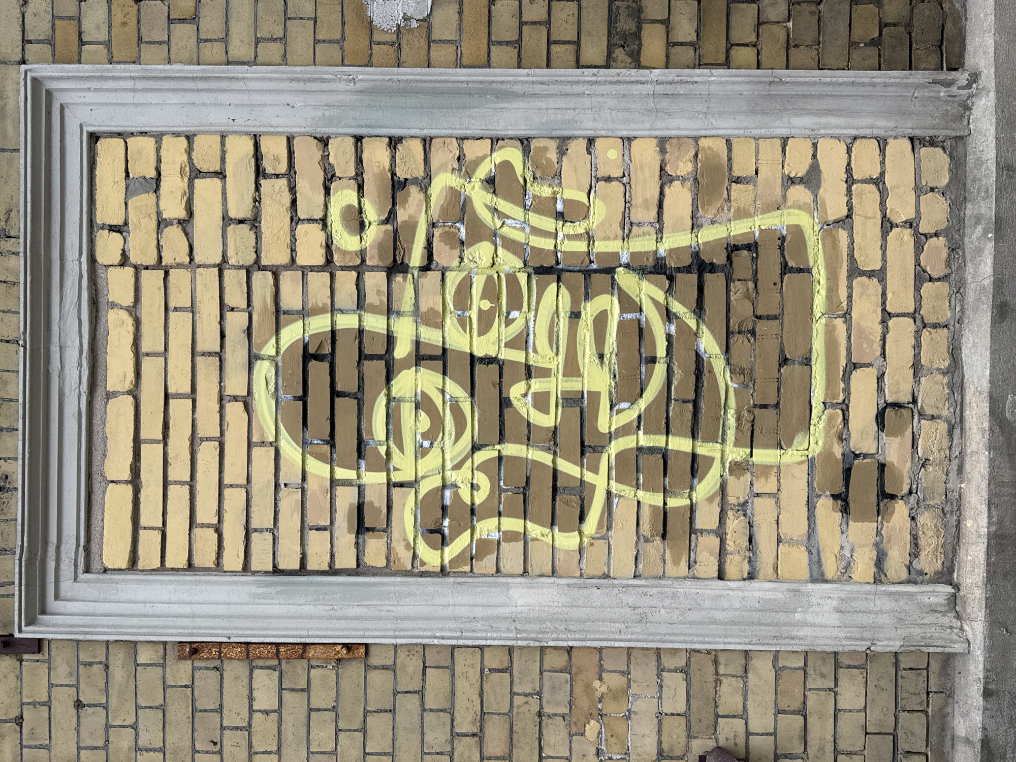 A rectangular bricked wall, framed by a concrete molding, features yellow graffiti. The graffiti consists of abstract lines and curves. The bricks are yellowish in color. The brick pattern extends beyond the concrete frame, both above and below. The top of the frame has a patch of white paint that is peeling off. The bottom of the frame has rusted metal pieces sticking out. The location is Riga, Latvia.