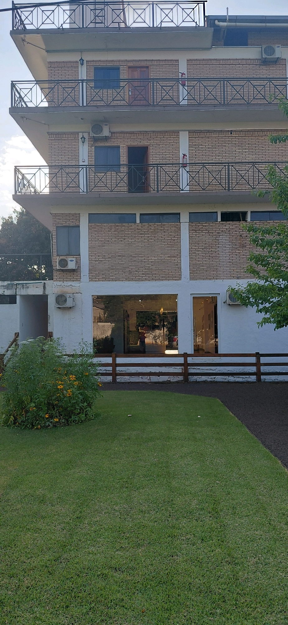 The image shows a multi-story building with a brick facade and balconies, located in Presidente Franco, Paraguay. The building has multiple windows and air conditioning units visible on its exterior. In front of the building, there is a well-maintained lawn, a small bush with yellow flowers, and a wooden fence. A person is visible through the large glass windows on the ground floor, appearing to be inside the building. The sky is partially cloudy.