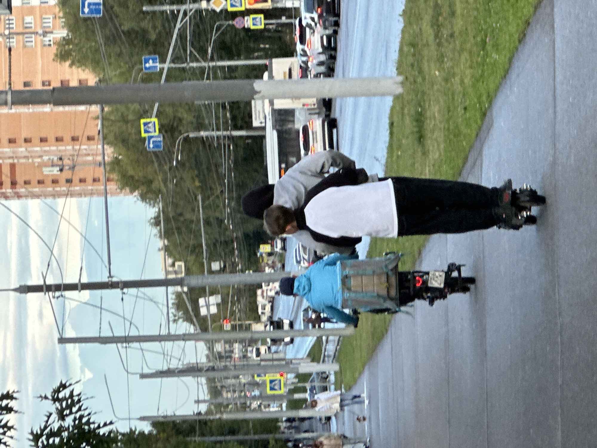 In Podolsk, Russia, on a cloudy day, two individuals are riding scooters on a sidewalk adjacent to a busy street. The person in the foreground is riding a black electric scooter, facing away from the camera, wearing black pants and a white t-shirt with black sleeves. Behind them, a second person, partially obscured, is riding a blue scooter with a large, gray insulated delivery box attached to the back. This scooter is also facing away from the camera. The street alongside the sidewalk is populated with several cars and a bus, indicating moderate traffic. Overhead, a complex network of utility wires and poles stretches across the scene, with traffic lights and directional signs visible. In the background, a tall, orange brick building and numerous green trees line the street, suggesting an urban environment. The sidewalk is paved with gray concrete and bordered by a strip of green grass.