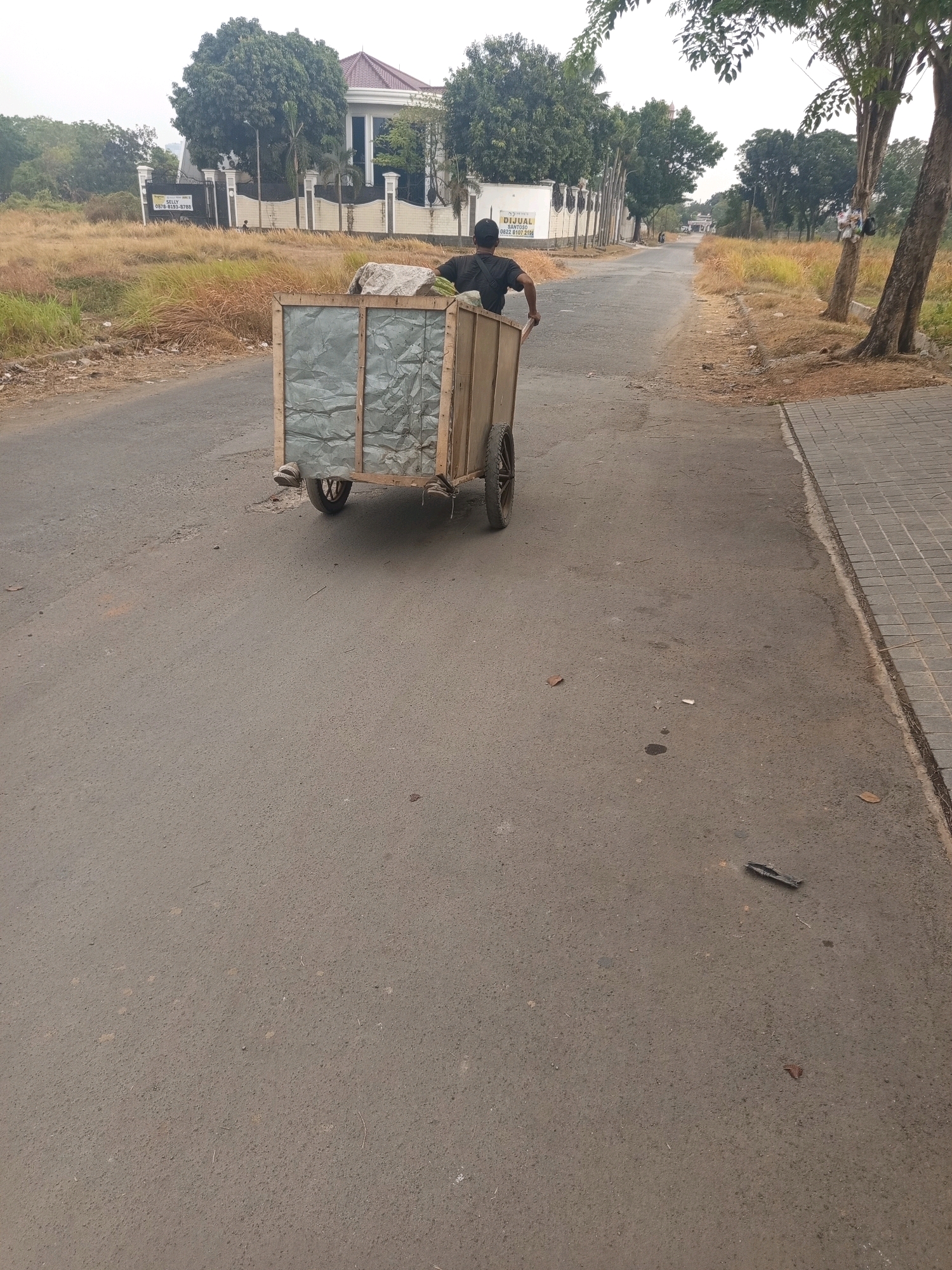 The image shows a street scene in West Jakarta, Indonesia. A man wearing a black t-shirt and a black baseball cap is pushing a wooden cart filled with what appears to be trash or recyclables. The cart has two wheels and a metal-lined interior. The man is walking away from the camera, down a paved road. To the left of the road, there is dry grass and a large house with a white fence and a sign that reads "DIJUAL" which means "FOR SALE" in Indonesian. To the right of the road, there are trees and a paved sidewalk. The sky is overcast.