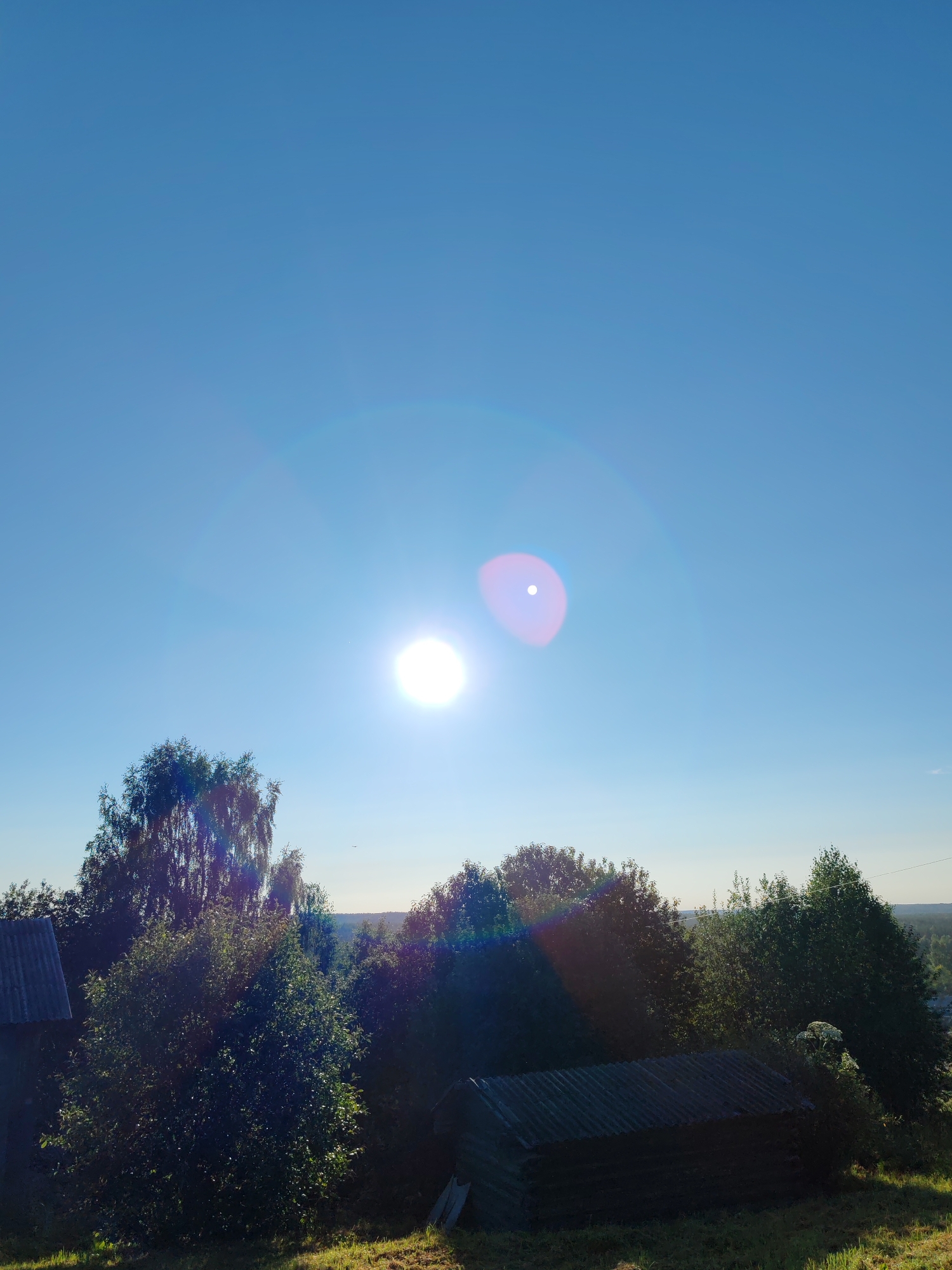 The image depicts a bright, sunny day in the rural settlement of Chukhlom, Russia, with a clear blue sky dominating the upper portion. The sun is visible as a luminous orb, causing lens flare with visible concentric circles of light. Below the sky, a dense line of lush green trees forms a natural horizon. In the foreground, there are elements of rural architecture, including a weathered, dark-colored shed with a corrugated roof, and a section of a grey, corrugated roof extending from a building on the left. The ground is covered in dry, green grass, suggesting a summer or early autumn setting. The overall impression is one of a peaceful, sun-drenched rural landscape.
