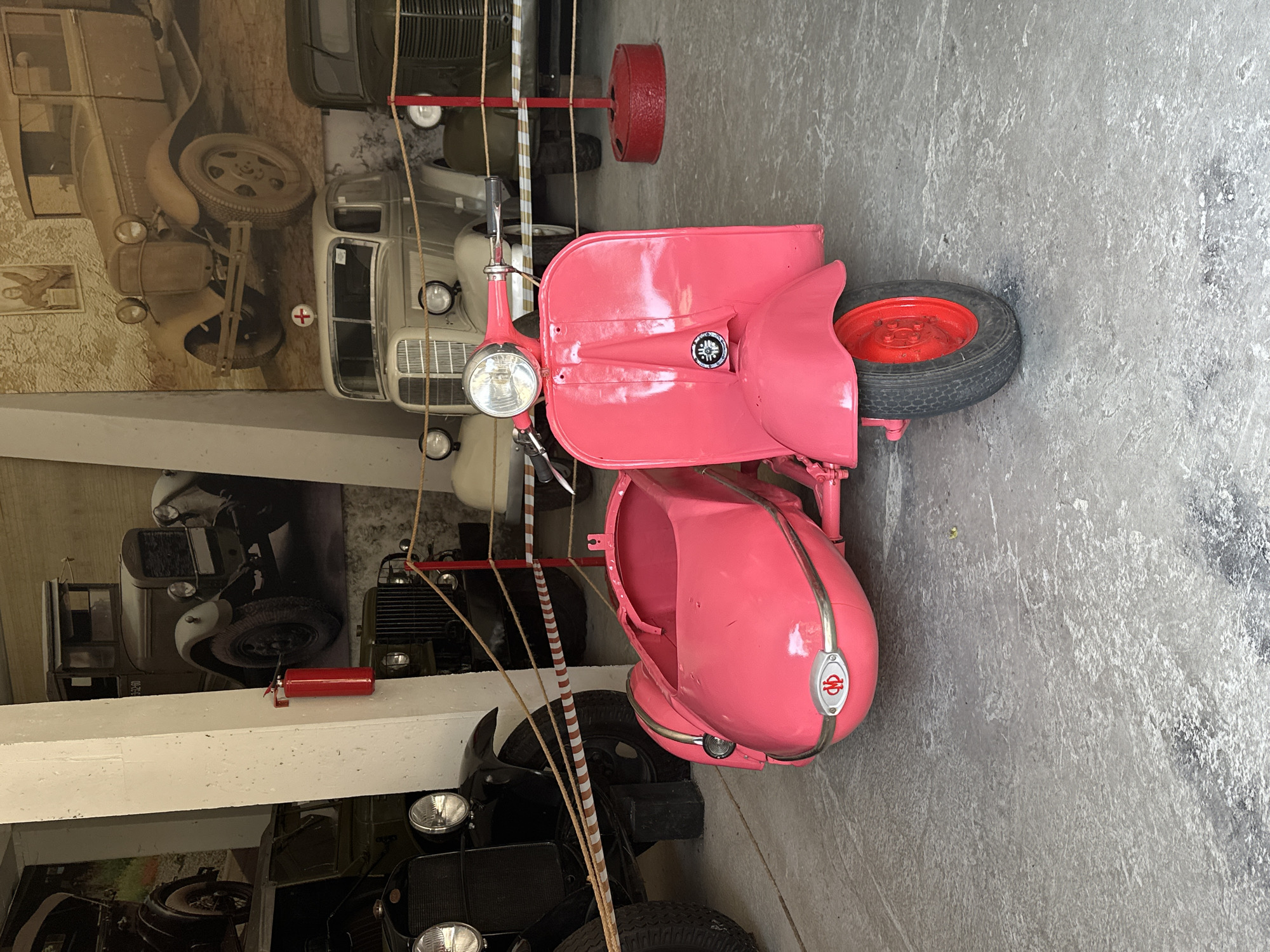 The photograph, taken in Sochi, Russia, depicts a pink vintage scooter with a sidecar displayed in a museum or exhibition hall. The scooter is positioned in the foreground, facing forward, with its handlebars, headlight, and front fender clearly visible. The sidecar, also pink, is attached to the right side of the scooter. Behind the scooter, several vintage vehicles are visible, including trucks and a white car with a red cross emblem. The scene appears to be indoors, with a concrete floor and large pillars supporting the ceiling. A red fire extinguisher is mounted on one of the pillars. The overall impression is one of a collection of historical vehicles on display.
