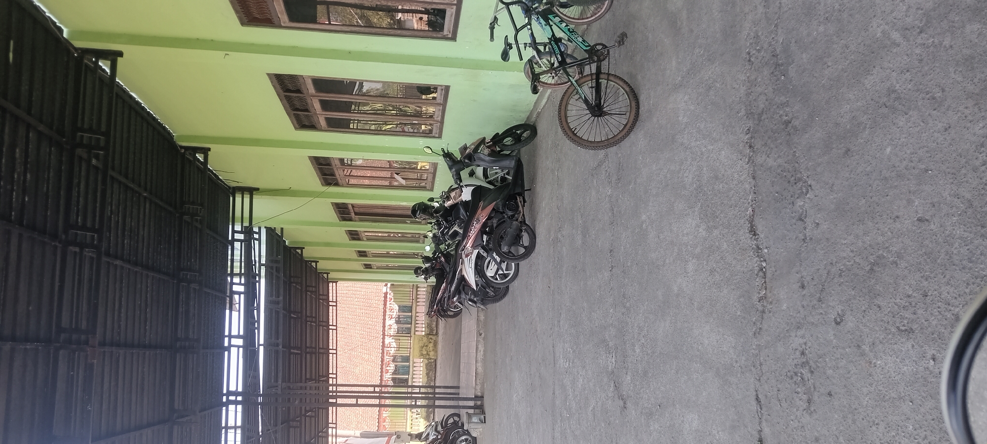 This image captures a street scene in Tempurejo, Indonesia, likely near a public building given the row of parked motorcycles and bicycles under a covered awning. The awning, constructed with dark metal beams and corrugated roofing, extends along the side of a building painted a light green. Several windows with wooden frames are visible on the building's exterior. In the foreground, a concrete surface shows some cracks. Parked motorcycles, mostly scooters, are lined up under the awning. To the right, two bicycles, one with a prominent red tire, are also parked. In the background, a building with a reddish-tiled roof and decorative elements can be seen, suggesting it might be a school or community center. The scene appears to be in a relatively open area, possibly a parking lot adjacent to a facility.