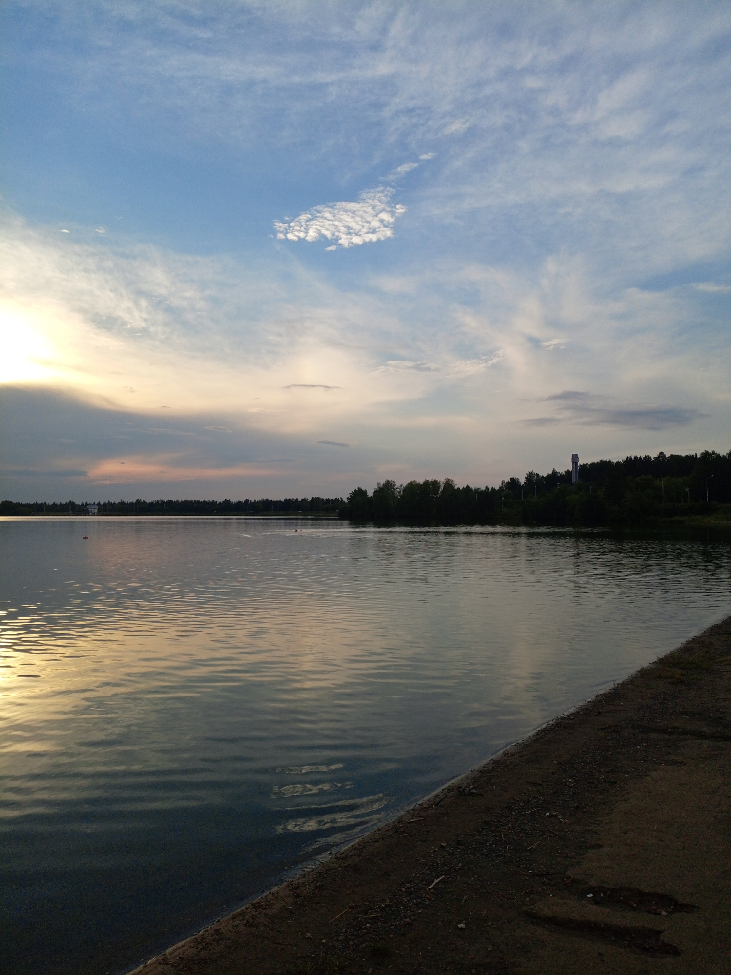 The image depicts a serene body of water, likely a lake or reservoir, with a wooded shoreline and a cloudy sky. The sun is setting or rising, casting a warm glow across the water and sky. In the distance, a tall, slender structure resembling a tower or monument stands silhouetted against the trees. The foreground shows a textured shore composed of sand and small stones. There are no people visible in the image. This scene is consistent with views of the landscape surrounding the city of Zheleznogorsk, Russia, particularly areas near water bodies that feature wooded shores and distinct man-made structures.