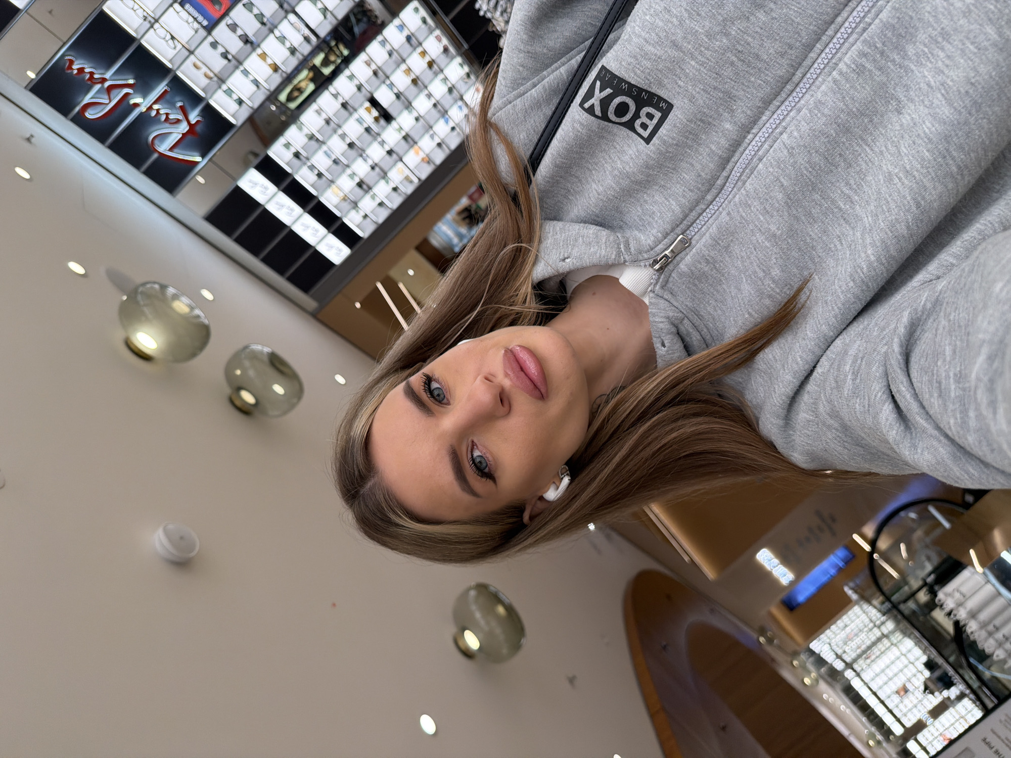 The image is a selfie taken in an indoor environment, likely a shopping mall or department store, as indicated by the visible retail displays. A woman with blonde hair is the primary subject; she is wearing a grey hooded sweatshirt with "BOX MENSWEAR" printed on it and has an earbud in her left ear. In the background, a Ray-Ban display suggests the location might be an eyewear store or a department store with an eyewear section. The lighting is bright, and the store fixtures include round, frosted glass light fixtures. A curved architectural element is visible in the lower right, potentially part of the store's design. The image was taken from a slightly low angle, as indicated by the upward tilt of the camera. The location is Berlin, Germany, because of the retail brands in the background.