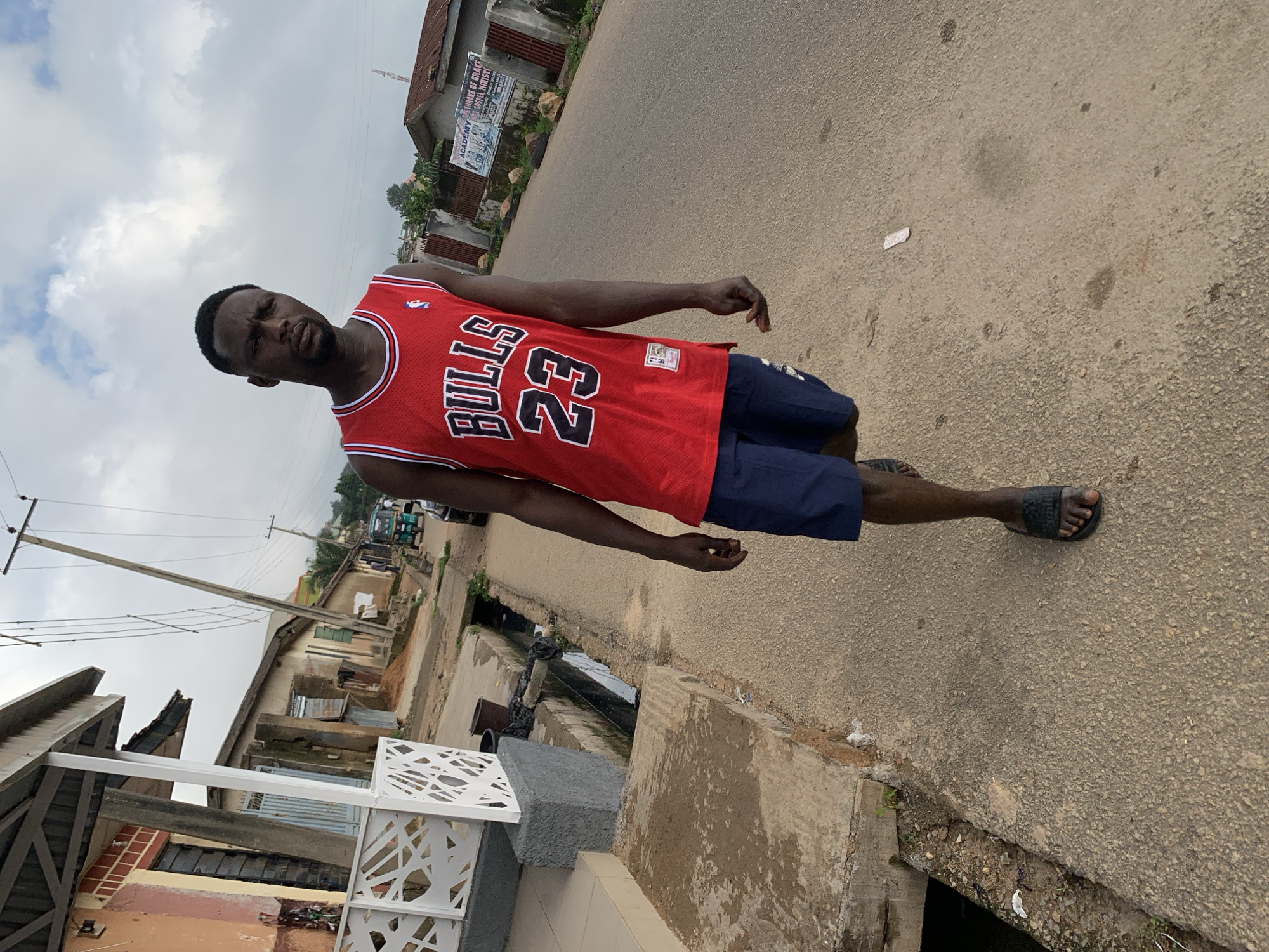 A young African man wearing a red Chicago Bulls basketball jersey with the number 23 and dark blue shorts walks on a paved sidewalk. He is looking towards the camera. The background shows a street scene with buildings, utility poles, a drainage ditch, and some vehicles. The environment appears to be a residential area in Jos, Nigeria. The sky is cloudy.