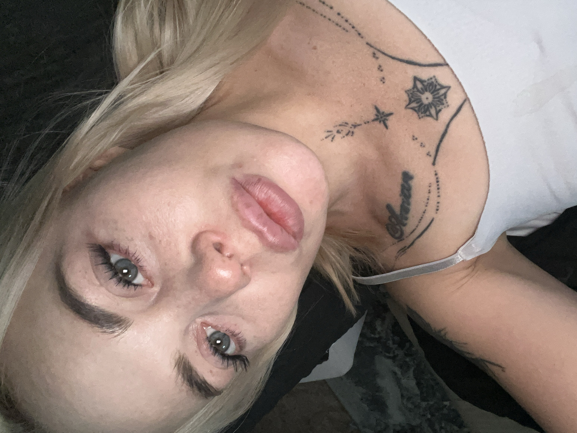 The image is a close-up, high-angle, indoor selfie of a young, Caucasian woman with blonde hair, pale skin, and light blue eyes. She is wearing a white tank top, and her face is angled towards the camera. She has prominent eyelashes and appears to have minimal makeup on. There are tattoos visible on her neck and shoulder. The background is dark and out of focus, with some textured material, possibly upholstery, visible on the left. The lighting is soft, creating a slightly reflective sheen on her skin and lips. The woman's expression is neutral, with her lips slightly parted. There are no other people or objects clearly identifiable in the scene. The location is not specified, but the indoor setting suggests a private space.