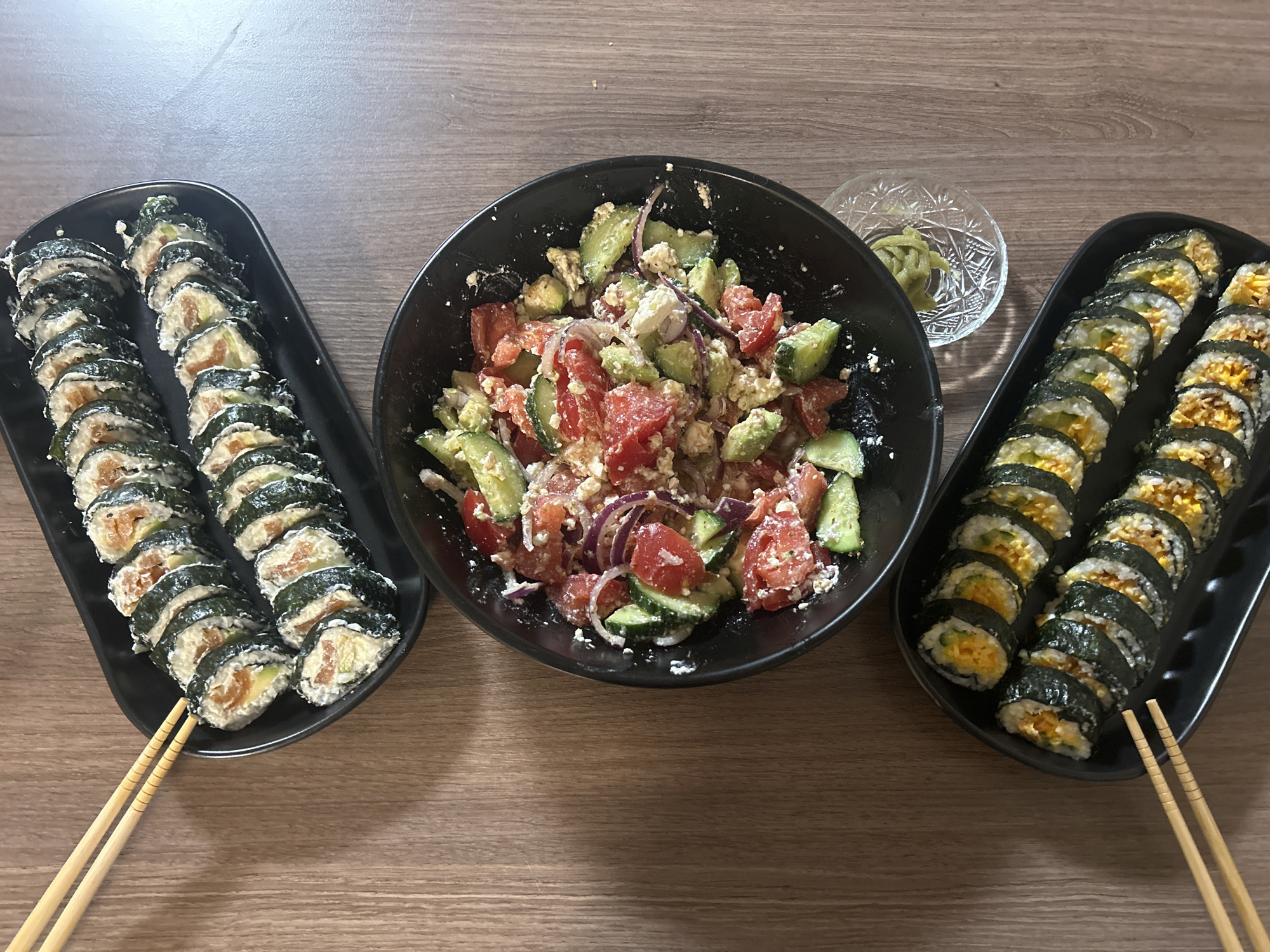 The image shows a meal consisting of two trays of kimbap, a bowl of salad, and a small dish of wasabi, arranged on a wooden table. The kimbap on the left tray appears to be filled with salmon, while the kimbap on the right tray contains a yellow filling. The salad in the black bowl consists of chopped tomatoes, cucumbers, red onions, and white cheese. A pair of chopsticks rests beside each tray of kimbap. The environment suggests an indoor setting, possibly a home. The image does not provide any information to determine a location in Veliko Tarnovo, Bulgaria.