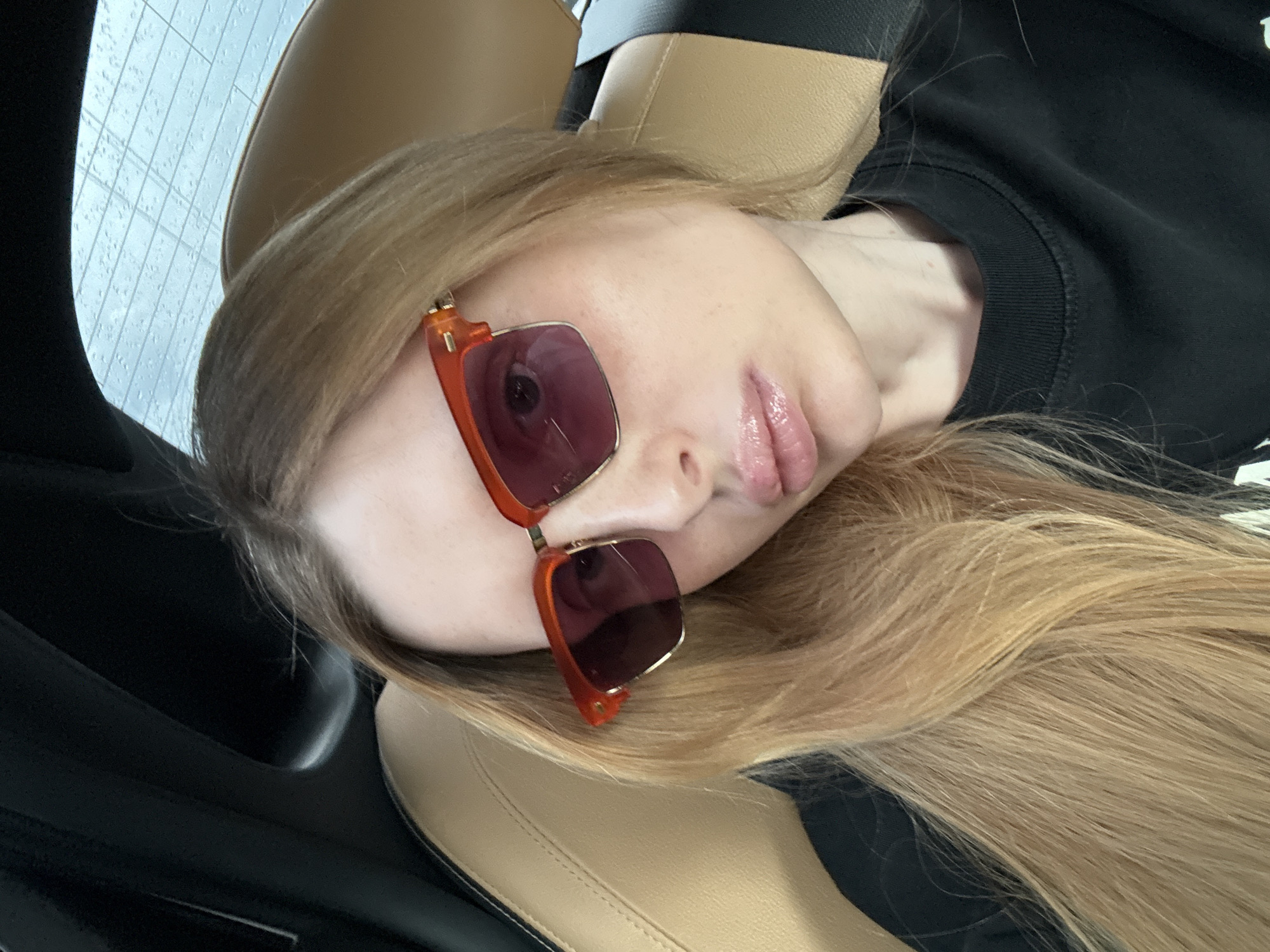 A woman with long, blonde hair is lying down, her head tilted to the side. She is wearing large, square sunglasses with orange frames and purple lenses. Her lips are parted slightly, and her eyes are not visible behind the sunglasses. She appears to be wearing a black sweatshirt. The setting seems to be inside a car, with tan leather seats visible in the foreground and a black interior on the left. The background shows a light blue surface with a grid pattern, possibly a window with rain on it. The lighting suggests it is daytime. The overall mood is casual and relaxed. There is no visible text in the image.