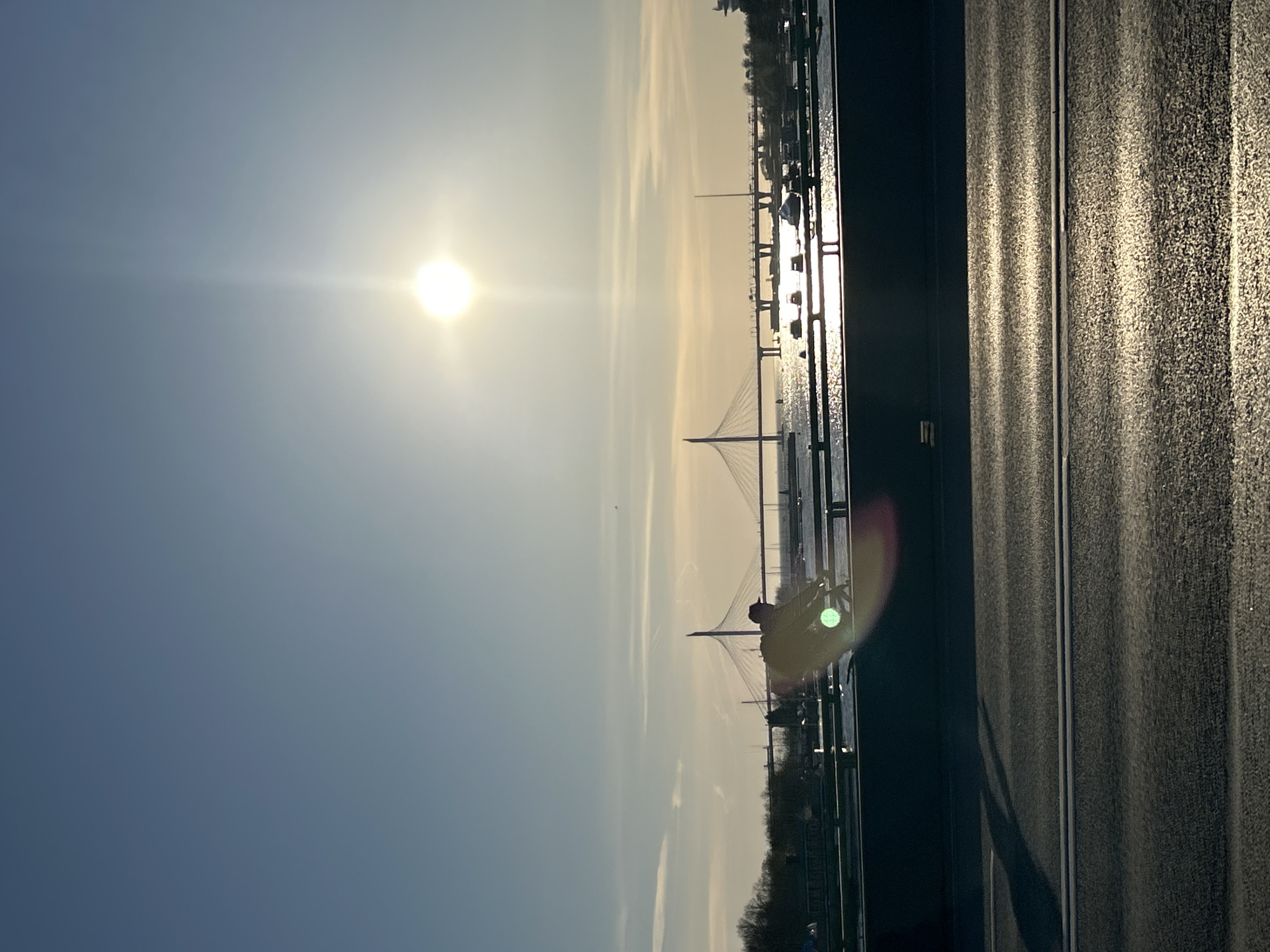 The image captures a view of Saint Petersburg, Russia, at sunrise or sunset, featuring the Cable-stayed bridge across the Petrovsky fairway. The sun, a bright orb, dominates the sky, casting a glare and obscuring some details. Below the sky, the bridge stretches across the water, its cables forming a distinctive pattern. Along the edge of the frame, to the right, is a roadway, with lane markings, as well as a fence. The overall tone of the image suggests a serene or tranquil setting.