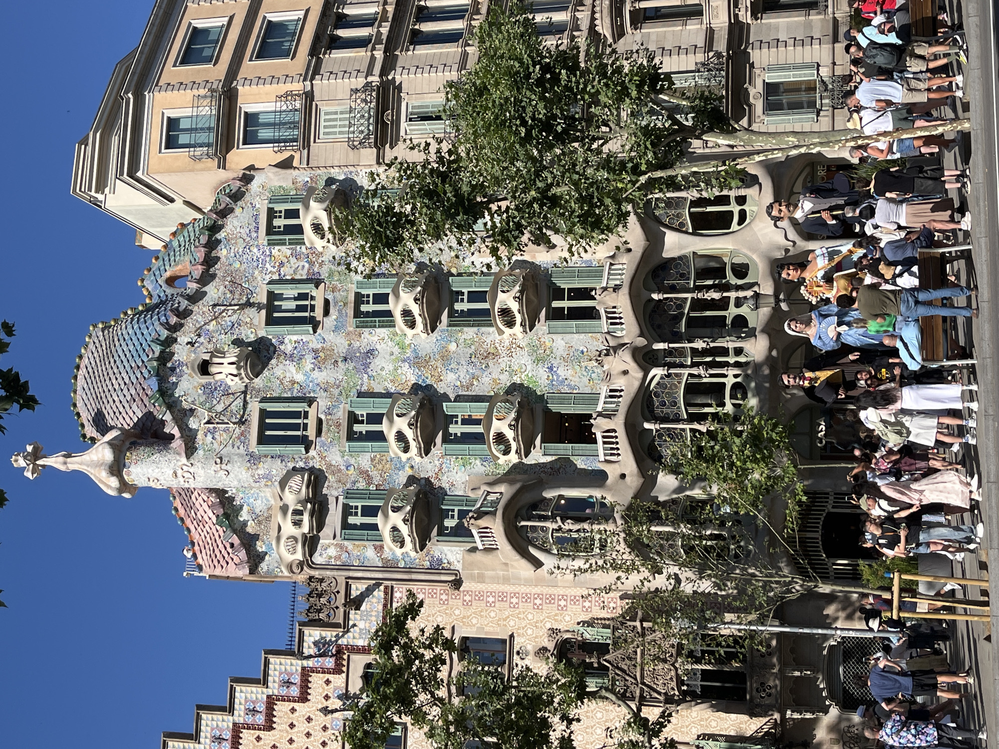 The image depicts Casa Batlló, a renowned building located on Passeig de Gràcia in Barcelona, Spain. The distinctive modernist architecture of Casa Batlló is characterized by its colorful mosaic facade, undulating organic shapes, and bone-like balconies. Adjacent to it, a more traditional building with a beige facade and balconies stands. In front of Casa Batlló, there is a crowd of tourists gathered. Among them are several large, costumed figures that appear to be part of a cultural display or performance, including a man in a suit and hat, a woman in a blue dress, and another woman in a yellow and red dress. Several trees with green foliage are strategically placed in the foreground and between the buildings, adding to the urban landscape. People are seen walking, standing, and observing the architecture and the costumed figures, with some taking photographs. The sky is clear and blue, indicating a sunny day.
