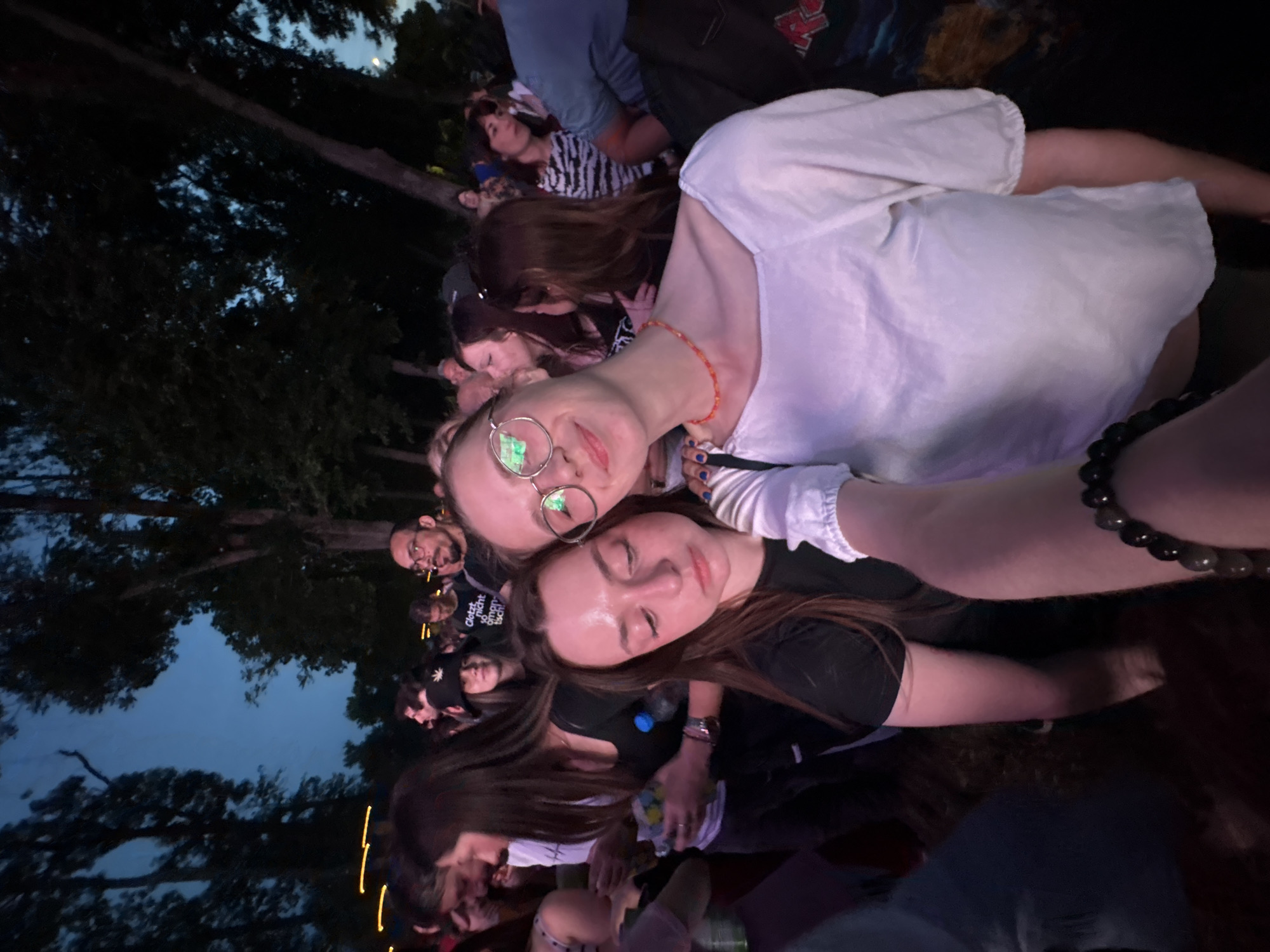 The image shows a selfie taken at a concert or outdoor event, likely in the evening. Two young women are in the foreground, posing for the camera. The woman on the left wears glasses and a light-colored top, while the woman on the right has long brown hair and wears a dark-colored top. Behind them, a crowd of people are visible. Trees surround the crowd and the sky is dark blue/grey. The event appears to be taking place outdoors, possibly in a park or a large open space. The location is Kragujevac, Serbia, though this is not visually evident.