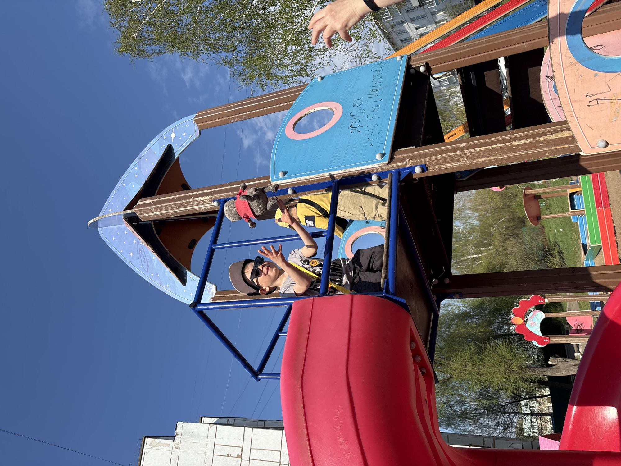 The image depicts a child wearing a hat and sunglasses inside a playground structure resembling a spaceship. The structure has a red slide and a blue control panel with a circular opening. Another child is visible above the first child, also inside the structure. The background shows a clear blue sky and a building. Based on the text on the blue panel, this scene takes place in the city of Chaikovsky, Russia.