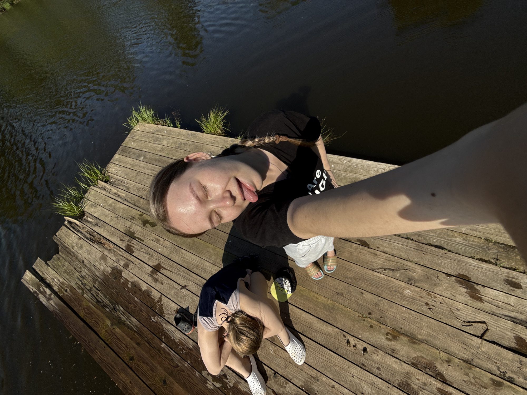 A high-angle selfie shows two light-skinned individuals on a wooden dock next to a body of water. The person taking the selfie is positioned at the top of the frame and to the right. They are sticking out their tongue and have their eyes closed. They are wearing a black shirt and light-colored shorts. To the left and slightly below them, the second person is sitting with their knees pulled up to their chest, facing away from the camera. They are wearing a two-toned top and white shoes. Between the two, there is a cap. The wooden dock has grass growing in the gaps between the boards. The water is dark and reflective. The location is Zhigalki, Russia.