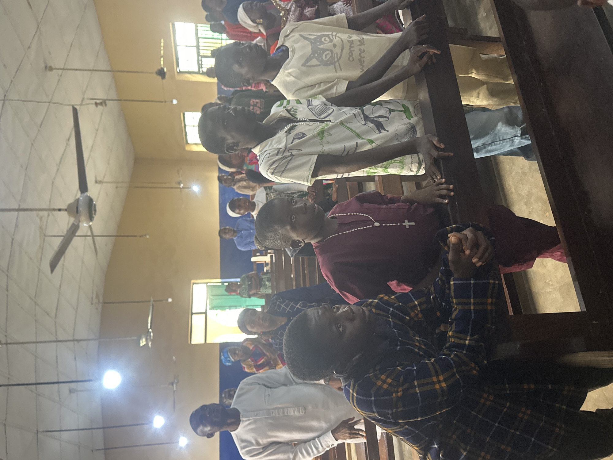 In Akwanga, Nigeria, a group of people, predominantly children and some adults, are gathered inside a building that appears to be a place of worship, possibly a church. The room has a high ceiling with fluorescent lighting and ceiling fans. The walls are painted a light yellow, and there are windows with green bars visible. The individuals are standing and are positioned behind wooden pews, with their hands resting on the pews.
In the foreground, a young boy wearing a maroon tunic and a beaded necklace with a cross is looking towards the right. Next to him, another boy in a plaid shirt is also looking to the right, with his hands clasped. To their right, two young boys are standing. One is wearing a white shirt with a green and white pattern, and the other is wearing a light yellow shirt with a cat graphic and text that reads "come we're led." Their hands are also on the pews.
In the background, adults and children are visible, some with head coverings. The overall atmosphere suggests a religious gathering or service. The lighting is somewhat dim, with the overhead lights casting a soft glow. The floor appears to be concrete.