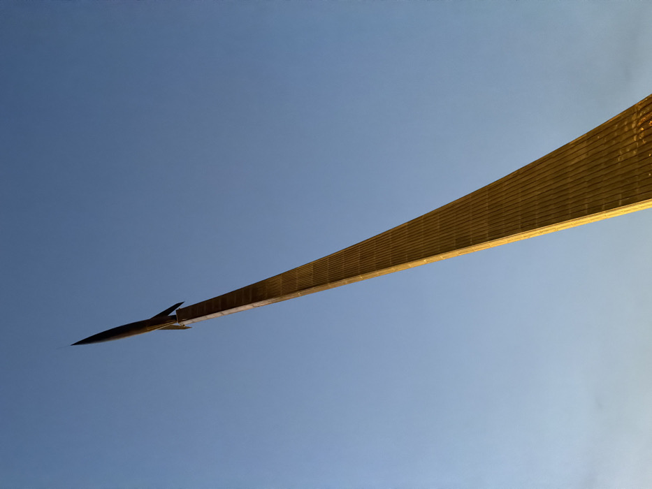 A low-angle, upward-looking photograph captures the Monument to the Conquerors of Space in Moscow, Russia, situated near the VDNKh exhibition center. The primary subject is a massive, tapering obelisk structure, clad in vertically arranged, reflective titanium panels that exhibit a warm, golden hue under the ambient light. A stylized rocket, resembling a Vostok launch vehicle, is prominently mounted at the apex of the obelisk. The monument extends diagonally from the lower right to the upper left quadrant of the frame, angled sharply skyward. The background consists entirely of a clear, gradient blue sky, lighter towards the implied horizon and deepening slightly towards the zenith, with no clouds visible. No individuals, additional subjects, or actions are depicted within the frame.