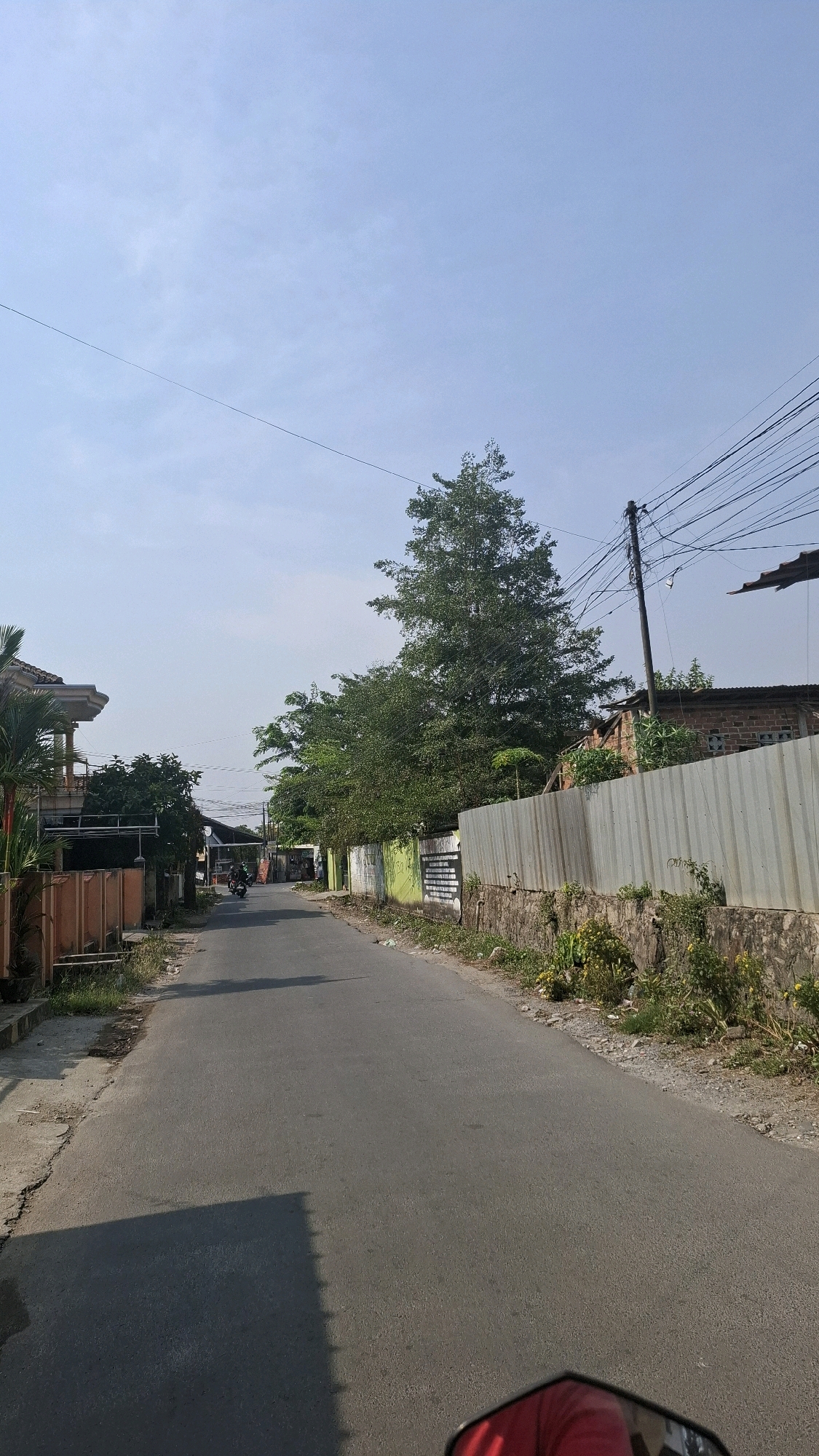 The image depicts a street scene in Bandar Lampung, Indonesia, under a clear blue sky. The road is asphalted and appears to be a local thoroughfare, with a slight incline. On the left side of the road, there are houses with modern architecture, some featuring balconies and orange-colored walls, accompanied by lush green plants and trees. Further down the road, a motorcycle is visible, with two individuals riding on it, heading away from the viewer. The right side of the road is lined with a metal fence, approximately six feet high, with a stone base. Behind the fence, there are residential buildings constructed with brick and corrugated metal roofing. Numerous power lines crisscross the sky, supported by a utility pole. The foreground shows the reflection of a person wearing a red shirt in what appears to be a motorcycle's rearview mirror. The overall environment suggests a residential area within the city, characterized by a mix of traditional and modern infrastructure, with evident vegetation and a sunny atmosphere.