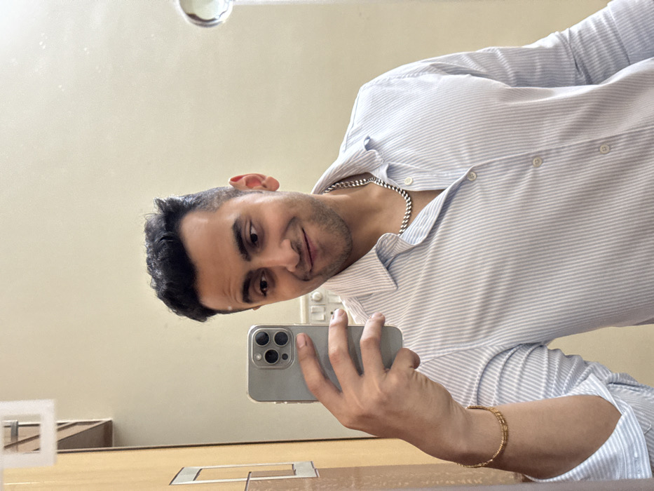 A man wearing a striped dress shirt and a silver chain necklace is taking a selfie in a mirror. He's holding a gray phone in his left hand, a gold bracelet adorns his wrist. The background is a neutral beige color. He has short dark hair and is smiling slightly. A small white object is visible in the lower left corner of the frame. A light fixture is seen in the upper center of the mirror.