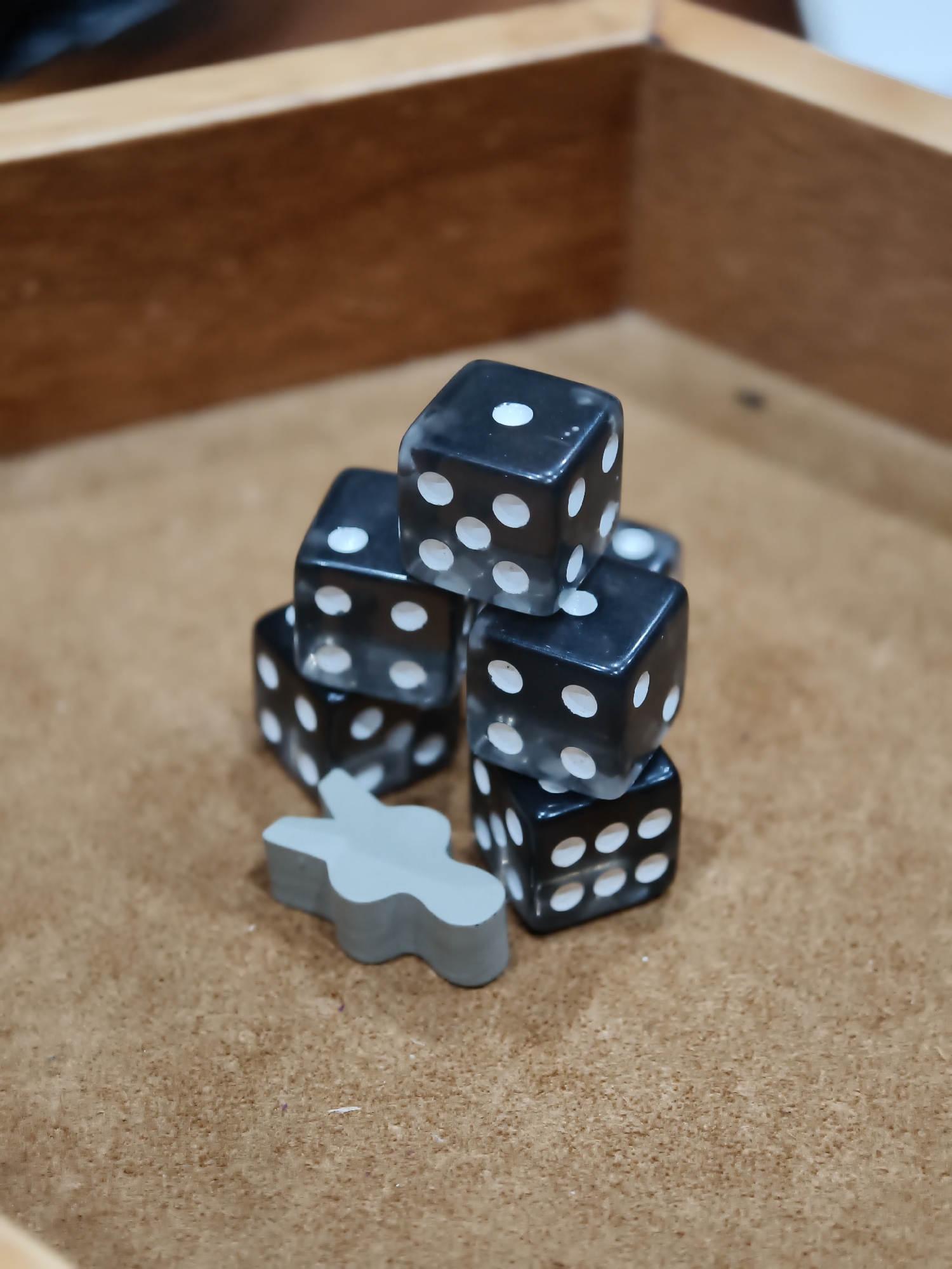 In a shallow, wooden game box, a stack of six black dice with white pips is arranged. The dice are piled in a pyramid shape, with two dice at the bottom, two in the middle, and two at the top. A single grey, human-shaped game token lies on the surface in front of the dice. The game box is placed on a brown, textured surface, possibly a table or the floor. The background consists of the upper portion of the wooden game box, showing its grain and construction. The lighting is soft and diffused, casting subtle shadows. The scene appears to be a still life setup, likely for a board game or a photograph related to tabletop gaming. There are no visible people or any discernible signs of the city of Đà Nẵng, Vietnam.
