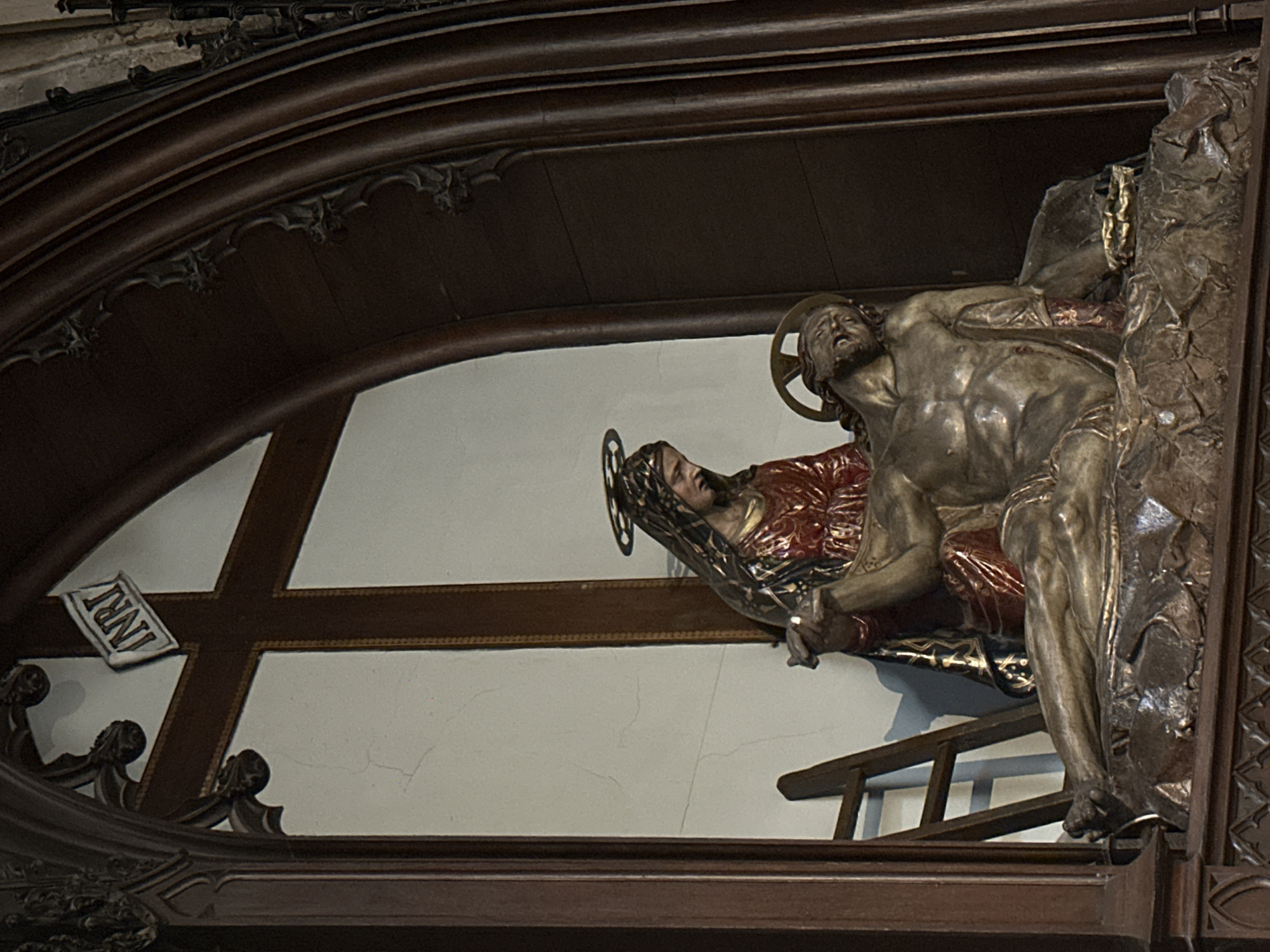 The image depicts a religious sculpture, likely a depiction of the Pietà, inside a church in Dieppe, France. The sculpture shows the Virgin Mary cradling the body of Jesus Christ after his crucifixion. Jesus' body is depicted lying across Mary's lap, with his head resting on her shoulder. Mary is shown with a sorrowful expression, her hands gently holding Jesus. Both figures have halos, indicating their divinity. A wooden ladder is visible to the left of the sculpture, leaning against a white wall. A wooden cross with the inscription "INRI" is positioned behind the sculpture, partially obscured by the religious figures and ornate wooden architectural elements that frame the scene. The overall setting suggests an altar or a devotional niche within a church.