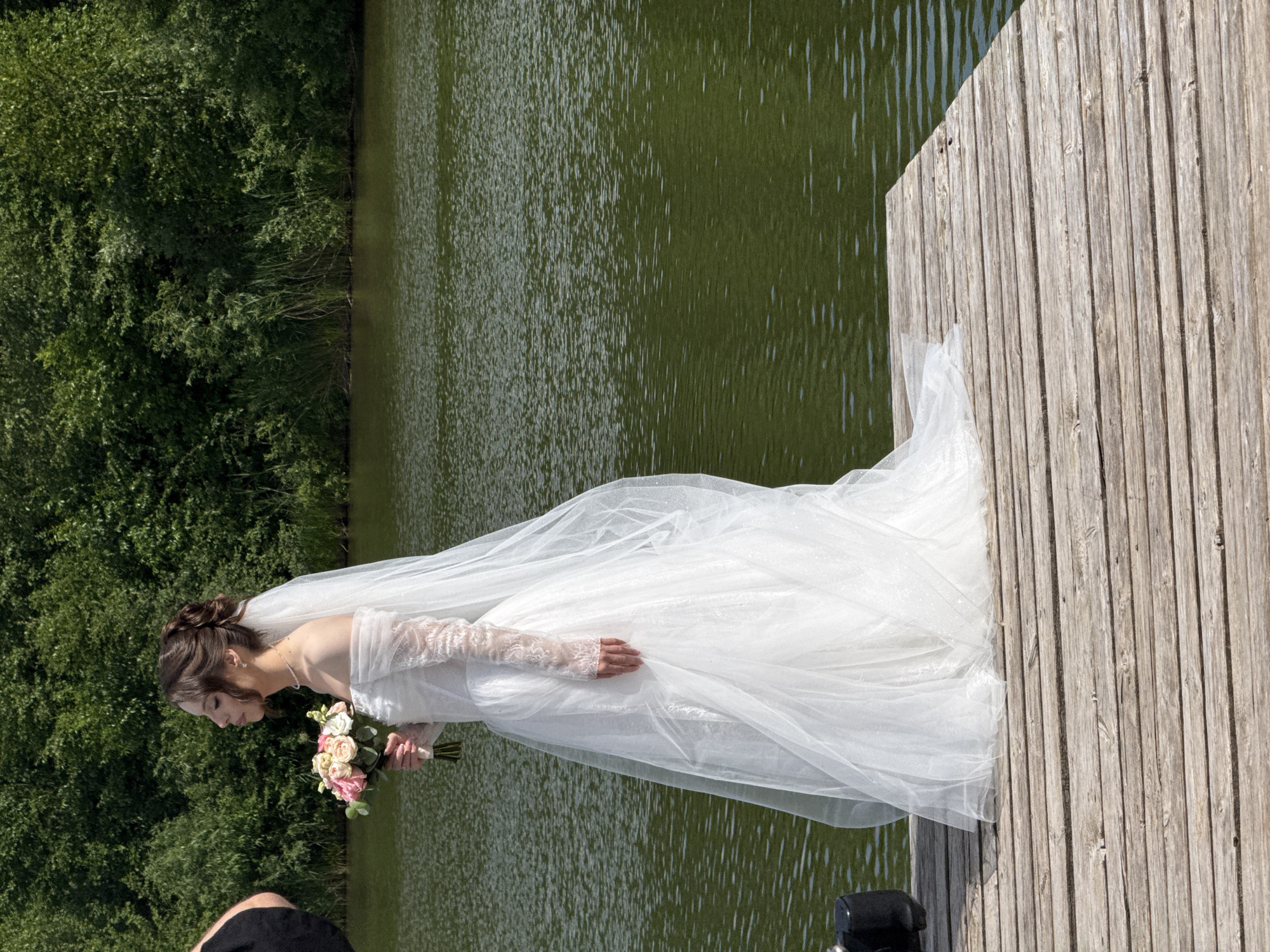 The image shows a bride in a white wedding dress standing on a wooden dock next to a body of water. The bride is holding a bouquet of flowers and has her eyes closed. The water is green and appears to be a lake or river. The background consists of lush green trees. It is a sunny day. The location is likely near the shore of Lake Votkinsk in Votkinsk, Russia, given the presence of a wooden dock and the natural, wooded landscape surrounding the water. The bride is the central subject, dressed in traditional bridal attire, suggesting a wedding ceremony or photoshoot.