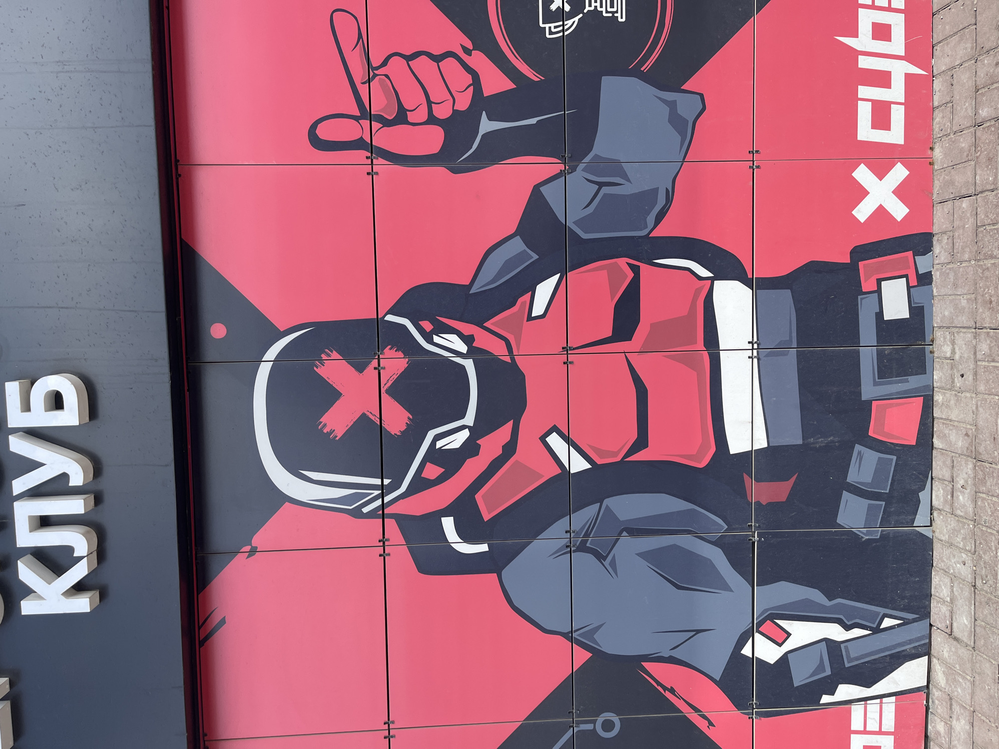 The original image is a vertically oriented photograph displaying a large, colorful mural on the side of a building, composed of square panels.
When viewed upright (rotated 90 degrees counter-clockwise), the central subject is a stylized graphic illustration of a human-like figure, possibly a cyber-soldier or gamer, rendered in bold red, black, grey, and white. The figure wears a distinct helmet featuring a prominent red "X" across the visor area and appears to be clad in tactical gear or armor. One hand is visible in the upper right portion of the mural, raised in a gesture that resembles either a "thumbs up" or a pointing motion. The background of the mural is predominantly red, accented with black and grey geometric shapes.
The activity depicted is the static display of this character as part of a building's facade, likely serving as branding or advertisement.
Notable details include the crisp, graphic art style, reminiscent of video games or comic books. The mural is outdoor, with a textured grey surface, possibly concrete or pavement, visible along the right edge of the original image. The lighting suggests it is daytime.
Visible text includes large, white, three-dimensional letters spelling "КЛУБ" (KLUB), meaning "CLUB" in English, positioned vertically on a black panel to the left of the mural. On the right side of the mural, white letters spelling "CYB" are vertically oriented, accompanied by a white "X" symbol. A small, stylized, and partially obscured symbol or text can also be seen within a black circular element near the top center of the mural. The location is Ulyanovsk, Russia.