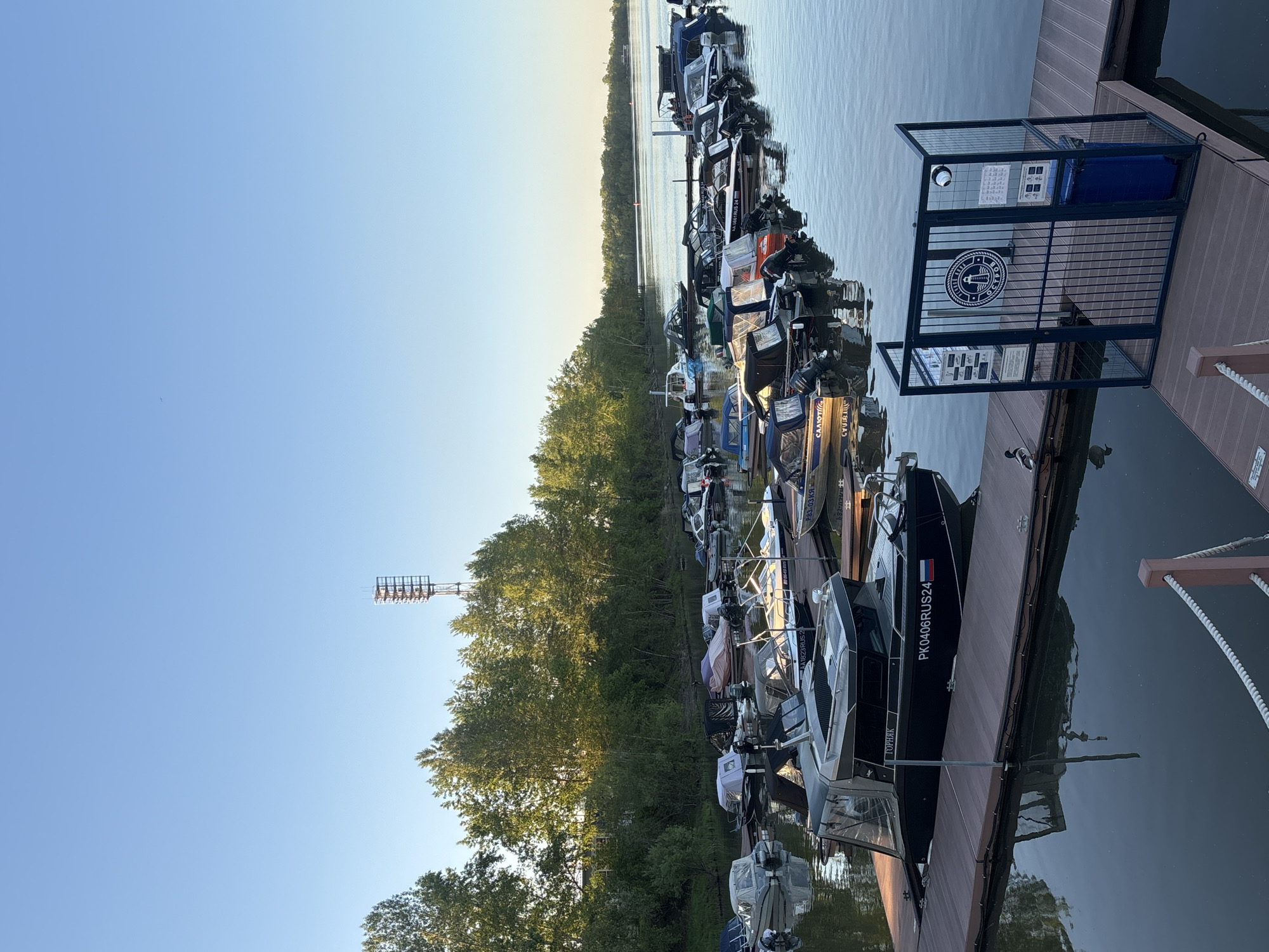 The image shows a marina located in Krasnoyarsk, Russia, filled with various recreational boats docked along a wooden pier. The boats are of different sizes and styles. The dock extends into calm water, with reflections visible on the surface. A forested area lines the opposite shore. An observation tower is visible amongst the trees. A blue sky is visible in the background. There is an enclosure with signage on the dock near the boats. One of the boats has the identification number "PK0406RUS24" displayed.